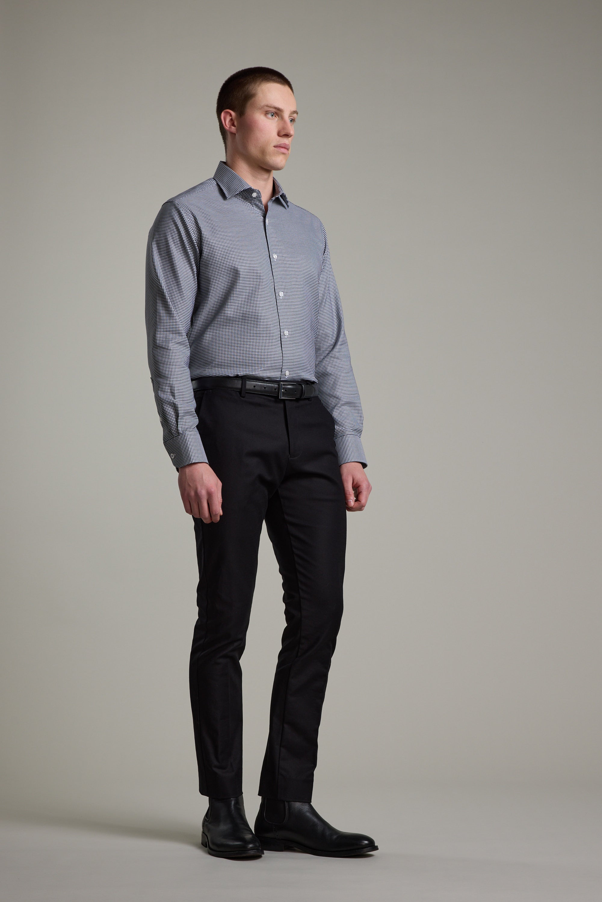 A man stands against a plain background wearing the Barkers Mitchel Check Business Shirt in a tailored fit, paired with black pants and dress shoes. He faces slightly sideways, arms relaxed and expression neutral.