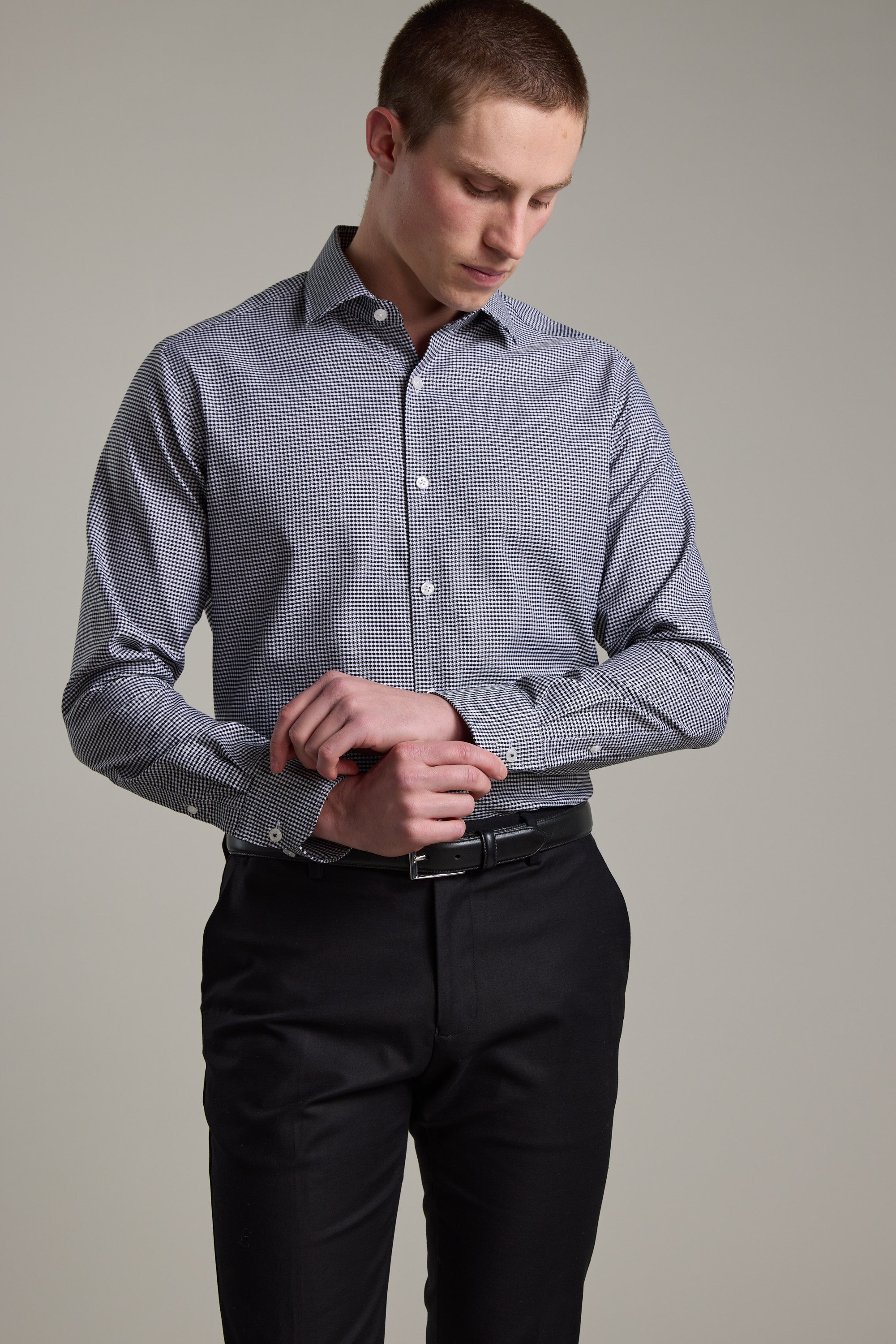 A man wearing the Barkers Mitchel Check Business Shirt and black trousers fastens his cuff while looking down, standing against a plain light gray background.