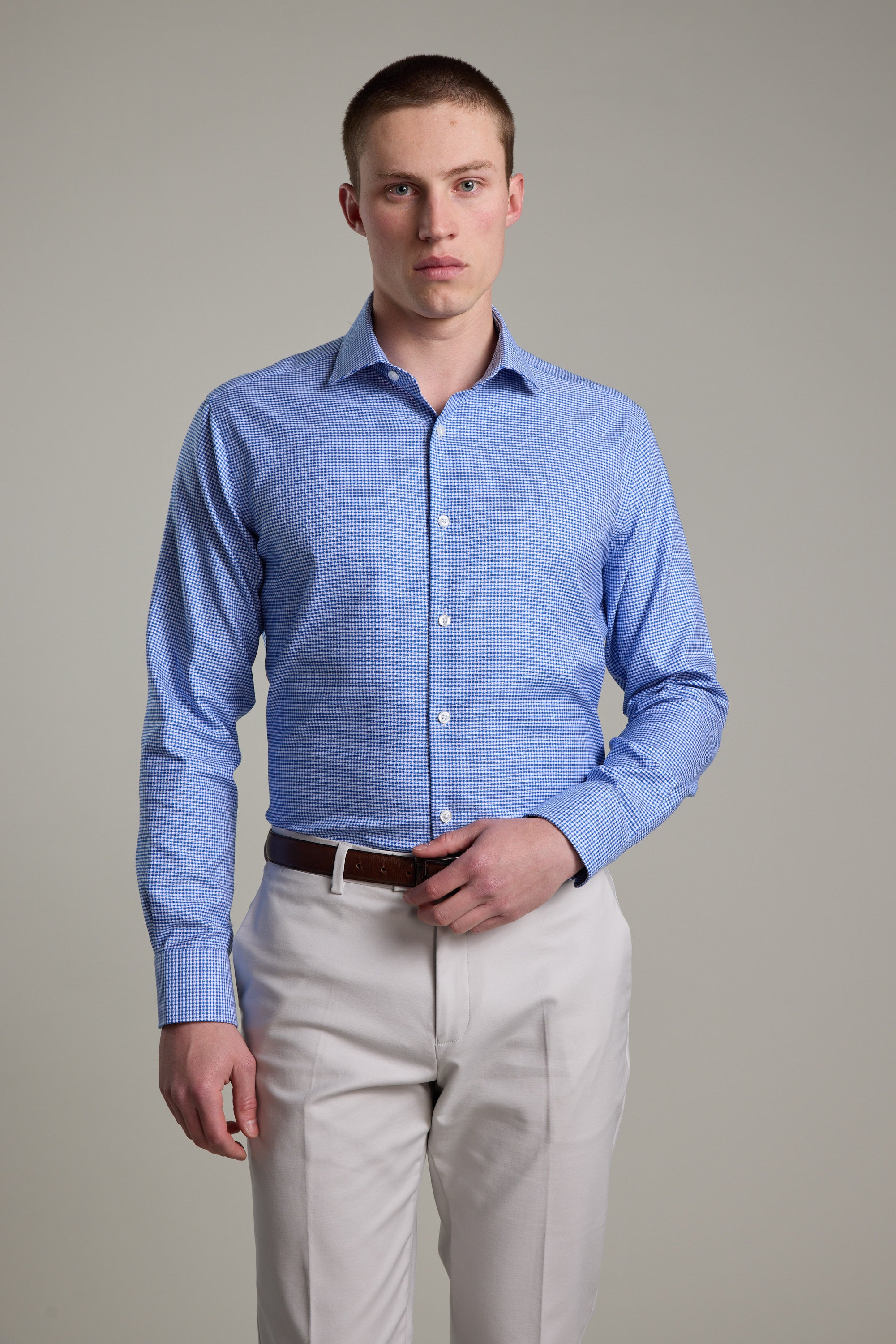 A man wearing the Barkers Mitchel Check Business Shirt, tailored fit with a light blue check pattern, is seen tucked into beige trousers with a brown belt, standing against a plain background and looking directly at the camera.