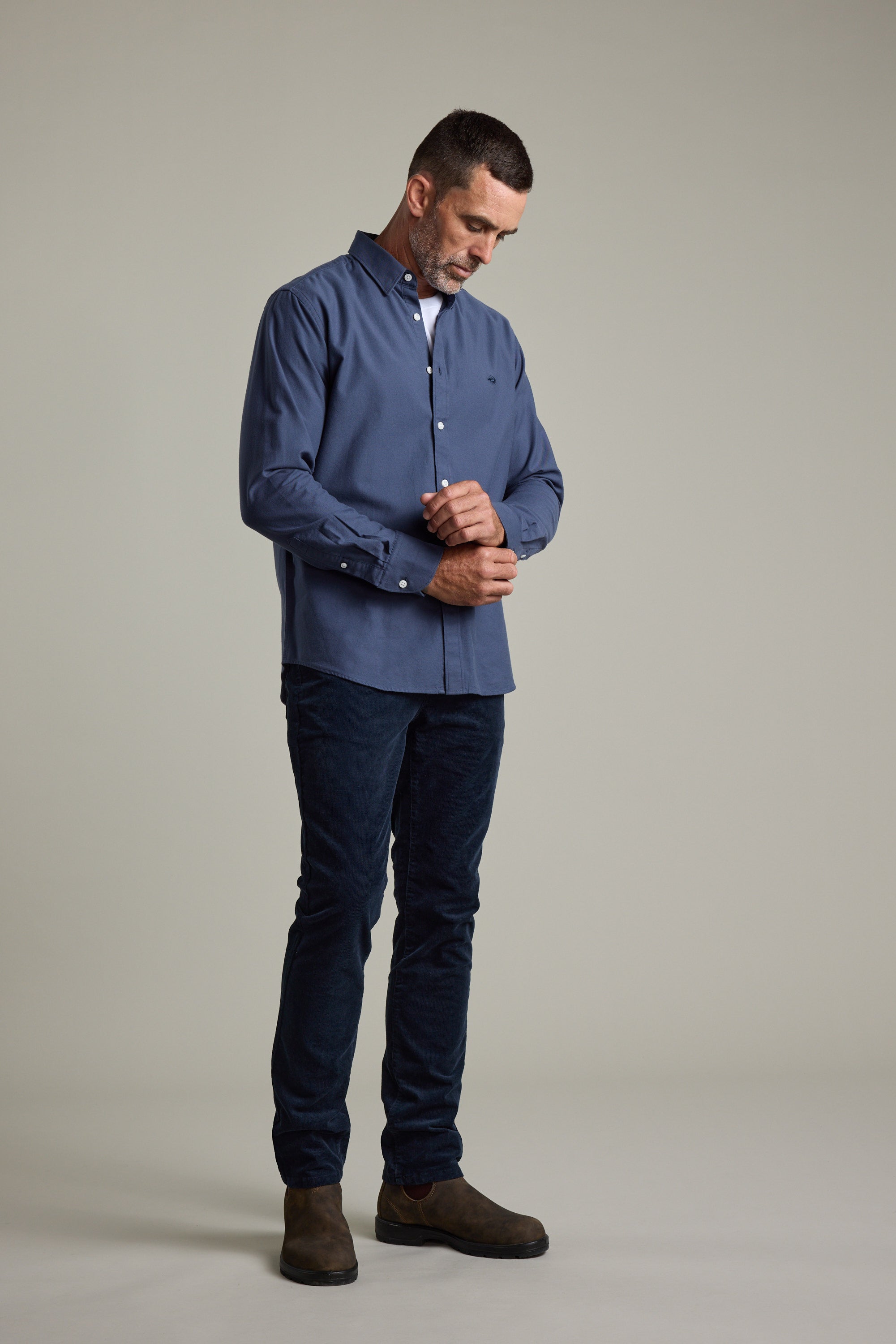 Man in Barkers Maynard Oxford Shirt and dark jeans adjusts his cuff on a plain background.