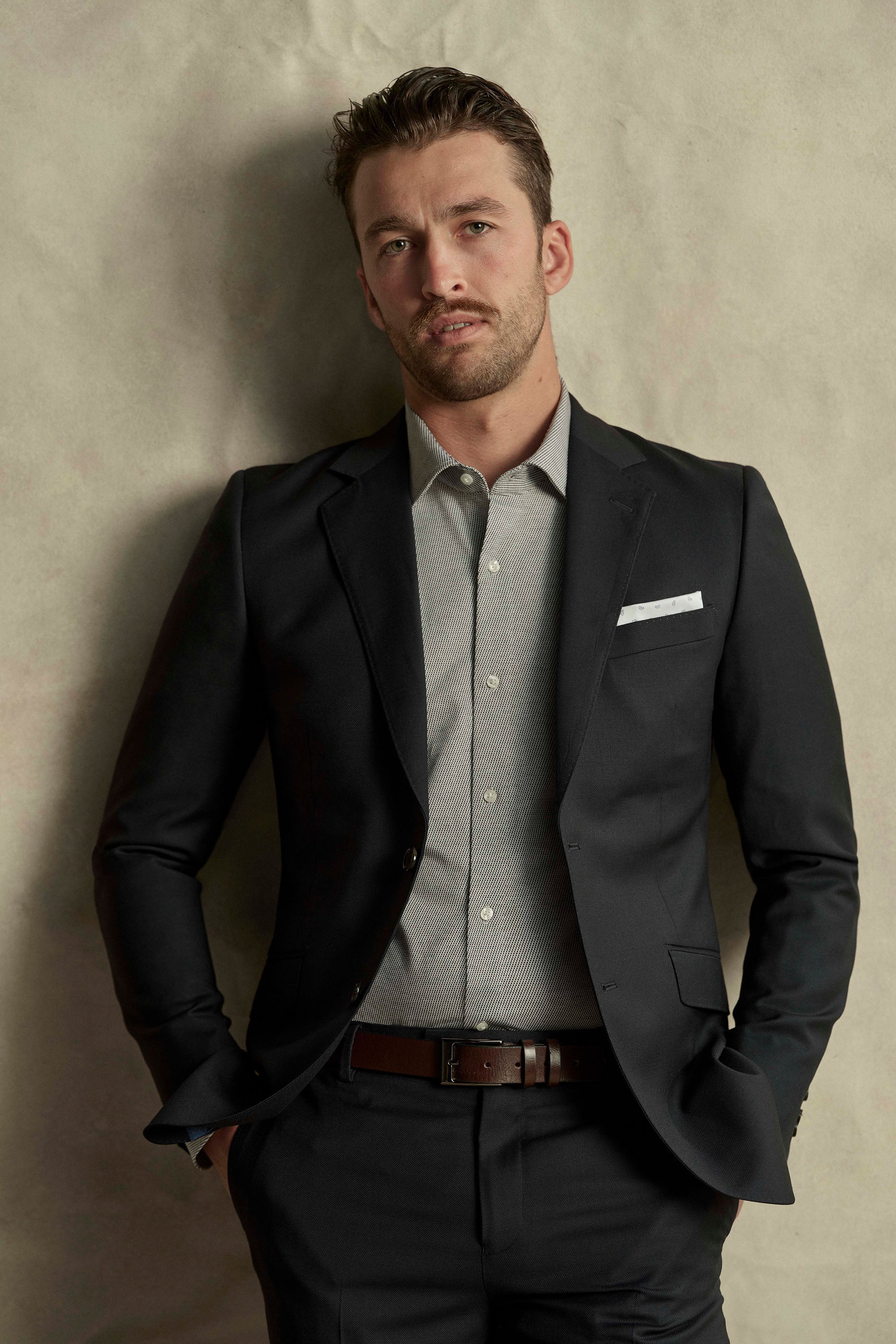 A man stands against a textured beige background, hands in pockets, wearing a Barkers Maclay Twill Blazer in black over a light gray dress shirt, looking at the camera with a neutral expression.