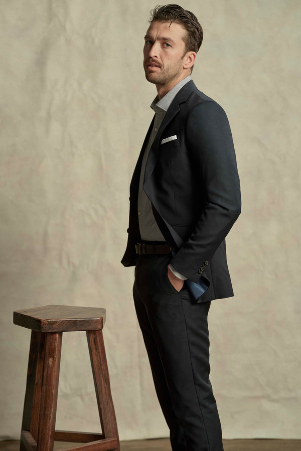 A man wearing the Barkers Maclay Twill Blazer over a light shirt with a pocket square stands beside a wooden stool, hands in pockets, posing against a neutral background.