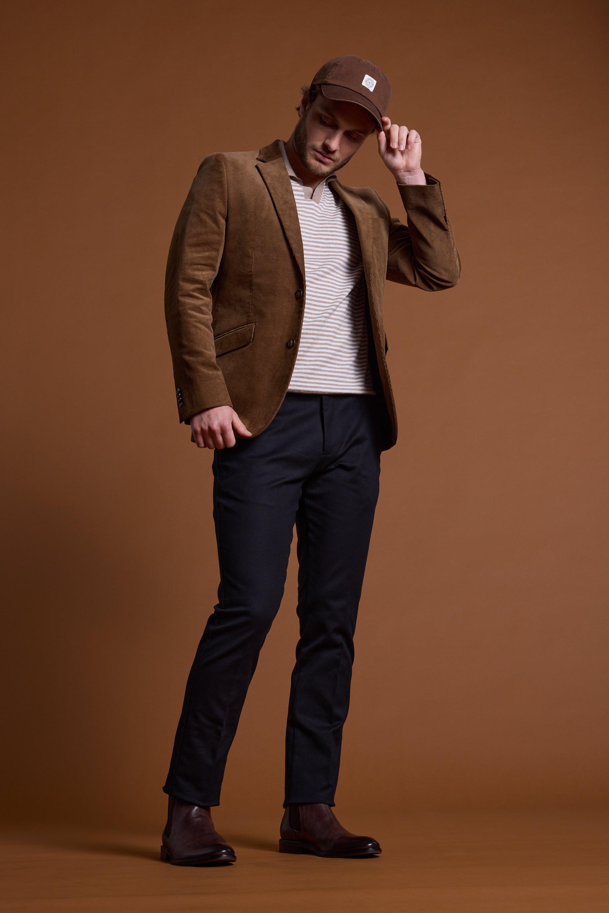 A man wears the Barkers Parkway Cotton Stripe Polo, paired with a brown blazer, black pants, brown shoes, and a brown cap. He stands against a brown background, tilting his cap forward while looking down.