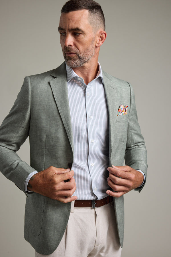 A man with short hair and a beard adjusts his Barkers Marion Texture Blazer, worn over a striped shirt, beige pants, and a brown belt—a smart choice for stylish summer occasions. He looks to the side while fixing his blazer with both hands.
