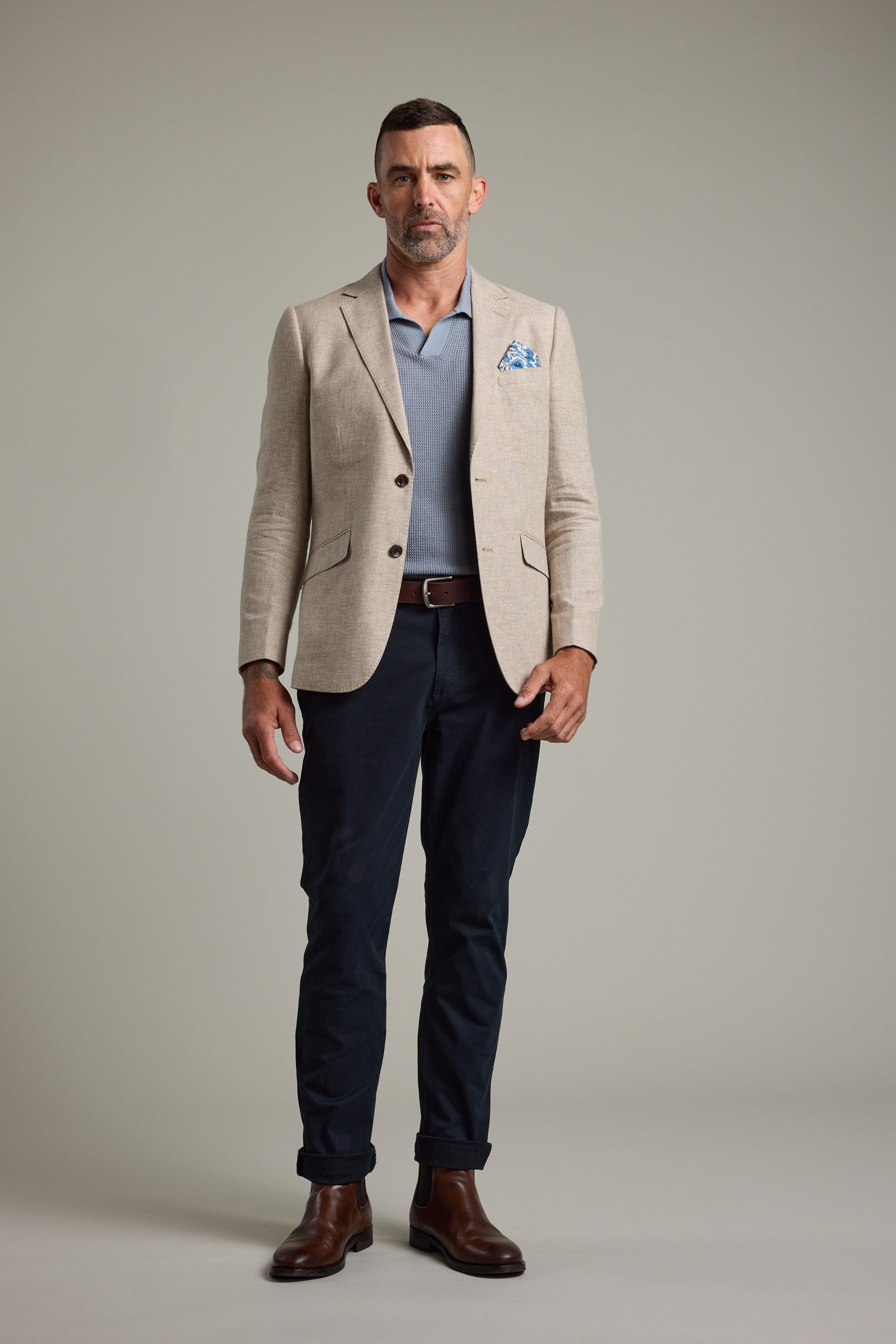 A man with short hair and a trimmed beard stands against a plain background, wearing Barkers' Welland Texture Blazer in beige with a blue pocket square, paired with a light blue polo shirt, dark blue pants, brown belt, and brown boots.