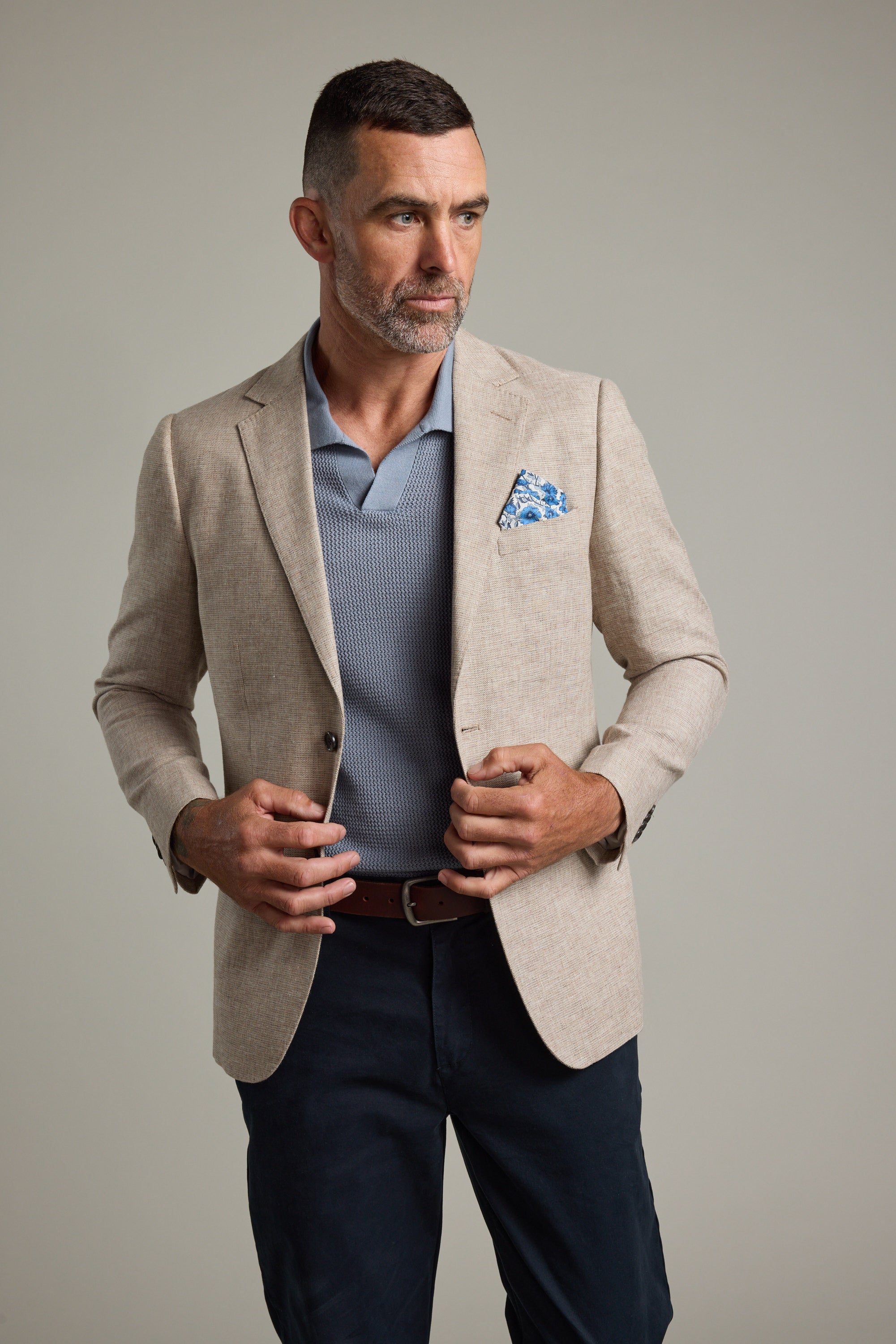 A man with short dark hair and a beard wears the Barkers Welland Texture Blazer in beige over a blue polo and dark pants, accessorized with a blue pocket square, looking to the side against a plain background.