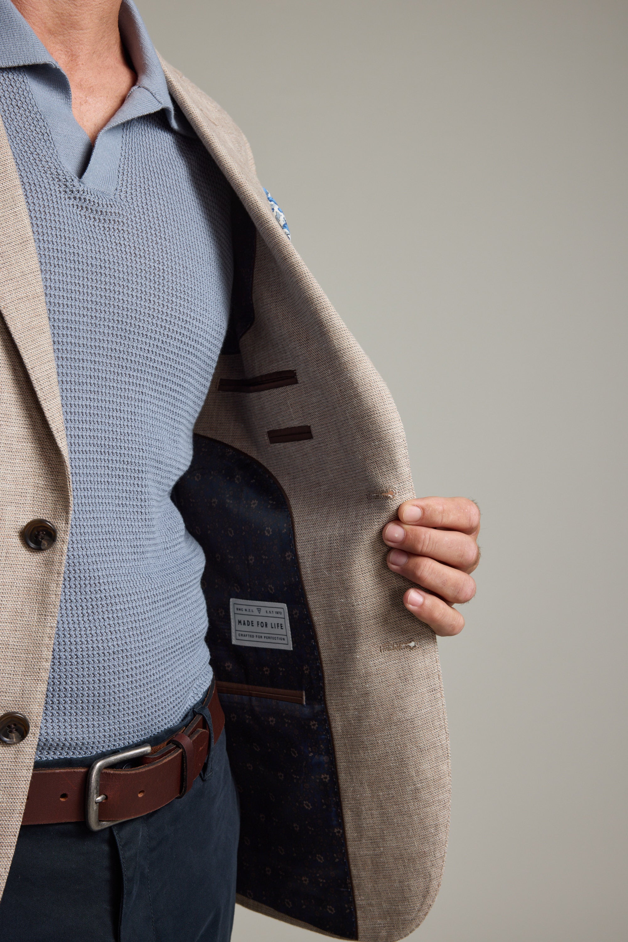 A person, face not visible, holds open the Barkers Welland Texture Blazer in beige linen to reveal its dark patterned lining and inner labels. They wear a light blue textured shirt, navy pants, and a brown belt.