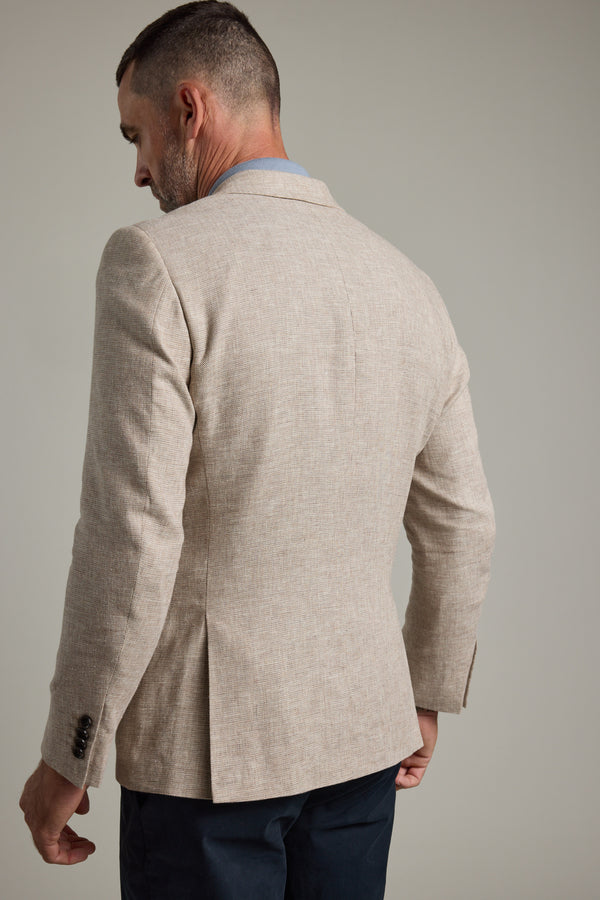 A man with short dark hair and a trimmed beard is seen from the back wearing the Barkers Welland Texture Blazer in light beige with dark sleeve buttons and dark trousers, standing against a plain light background.