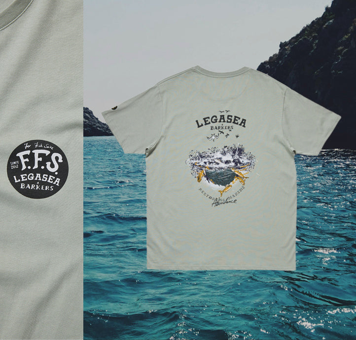 A pale gray t-shirt with a circular logo on the front reading “F.F.S LEGASEA BARKERS” and a large design on the back featuring ocean waves, birds, and text, is displayed against a background of blue ocean water and rocky cliffs.