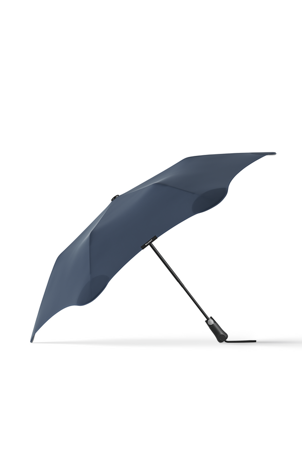 The Blunt Metro Umbrella by Blunt, featuring a dark blue, modern curved canopy and black handle, is open and angled upright on a white background.