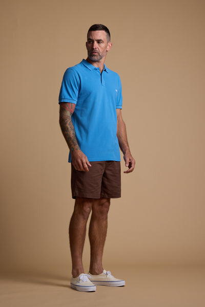 A man with short hair and tattoos on his left arm stands against a beige background, wearing a bright blue Barkers Regent Pique Polo in organic cotton, brown shorts, and white sneakers, with a neutral expression.