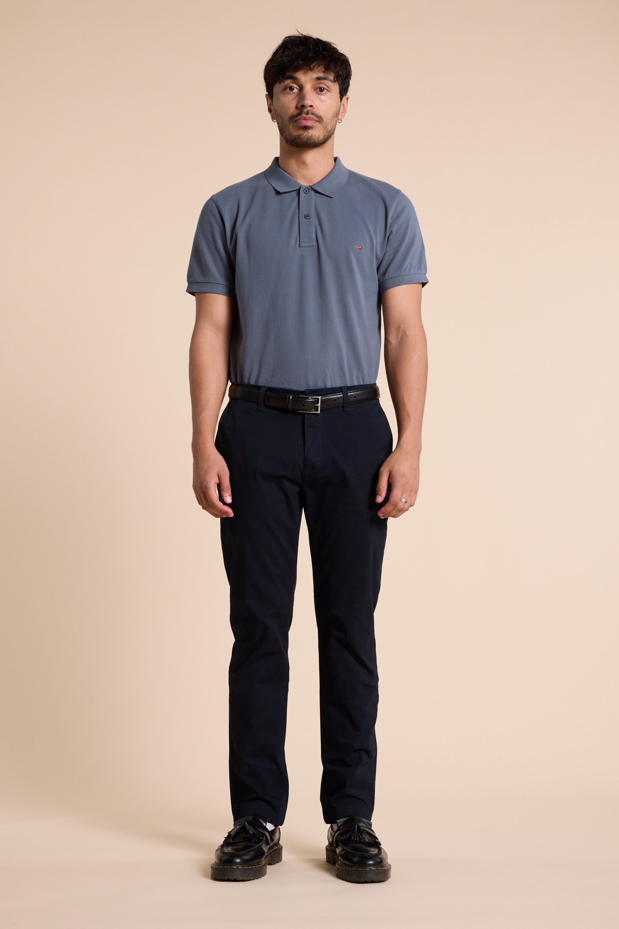 A man with short dark hair and a neutral expression stands facing forward against a plain beige background, wearing a blue Barkers Regent Pique Polo in organic cotton, dark pants, a black belt, and black shoes.