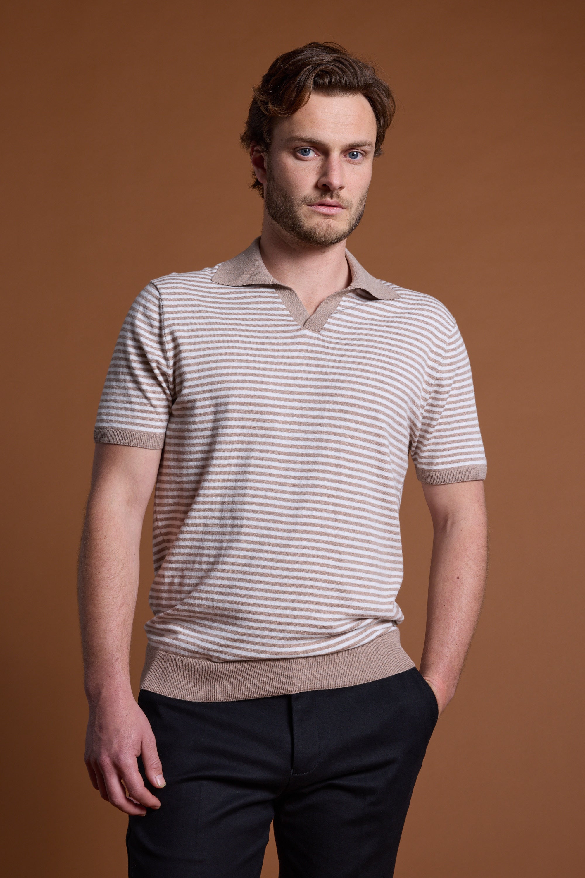 A man with light brown hair and a beard stands against a plain brown background, wearing Barkers’ Parkway Cotton Stripe Polo made from responsibly sourced cotton, dark pants, one hand in his pocket, and a neutral expression.