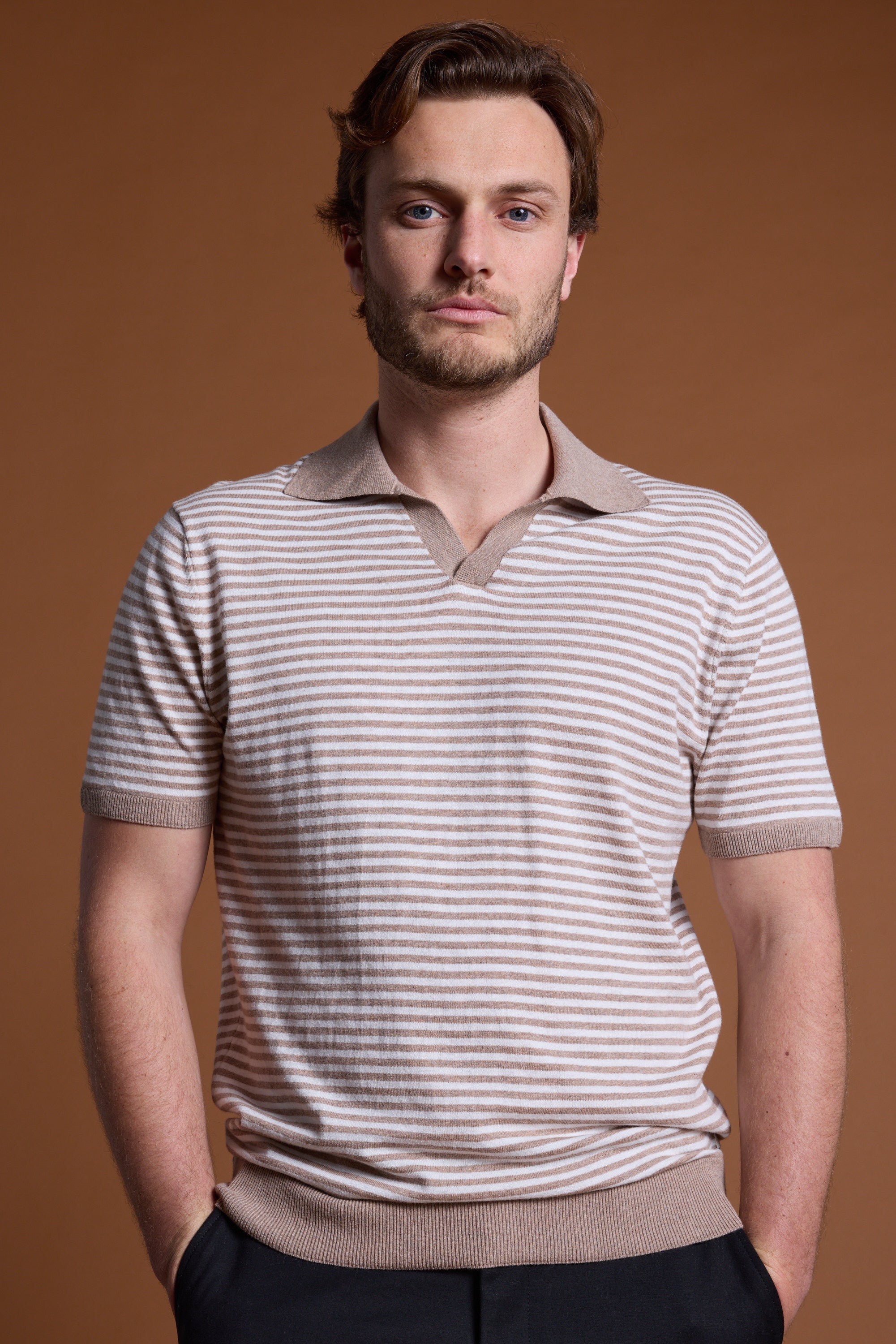 A man with light brown hair and a short beard stands against a brown background, wearing the Barkers Parkway Cotton Stripe Polo with a collar. His hands are in his pockets and he has a neutral expression.