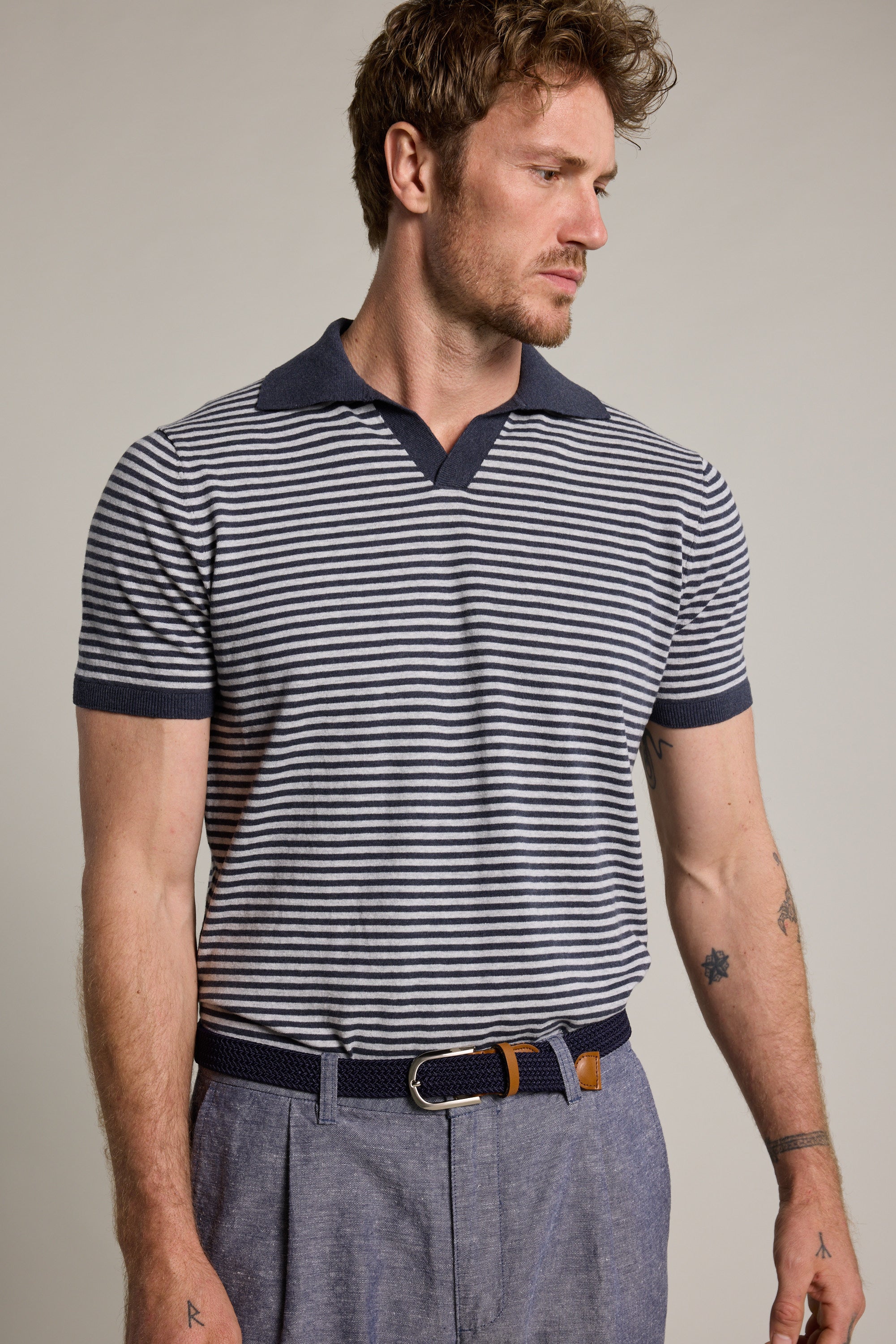 A man with short, wavy hair and facial hair models the Barkers Parkway Cotton Stripe Polo in navy and white, paired with light blue trousers and a navy woven belt. He looks left against a plain background, highlighting the lightweight polo shirt.