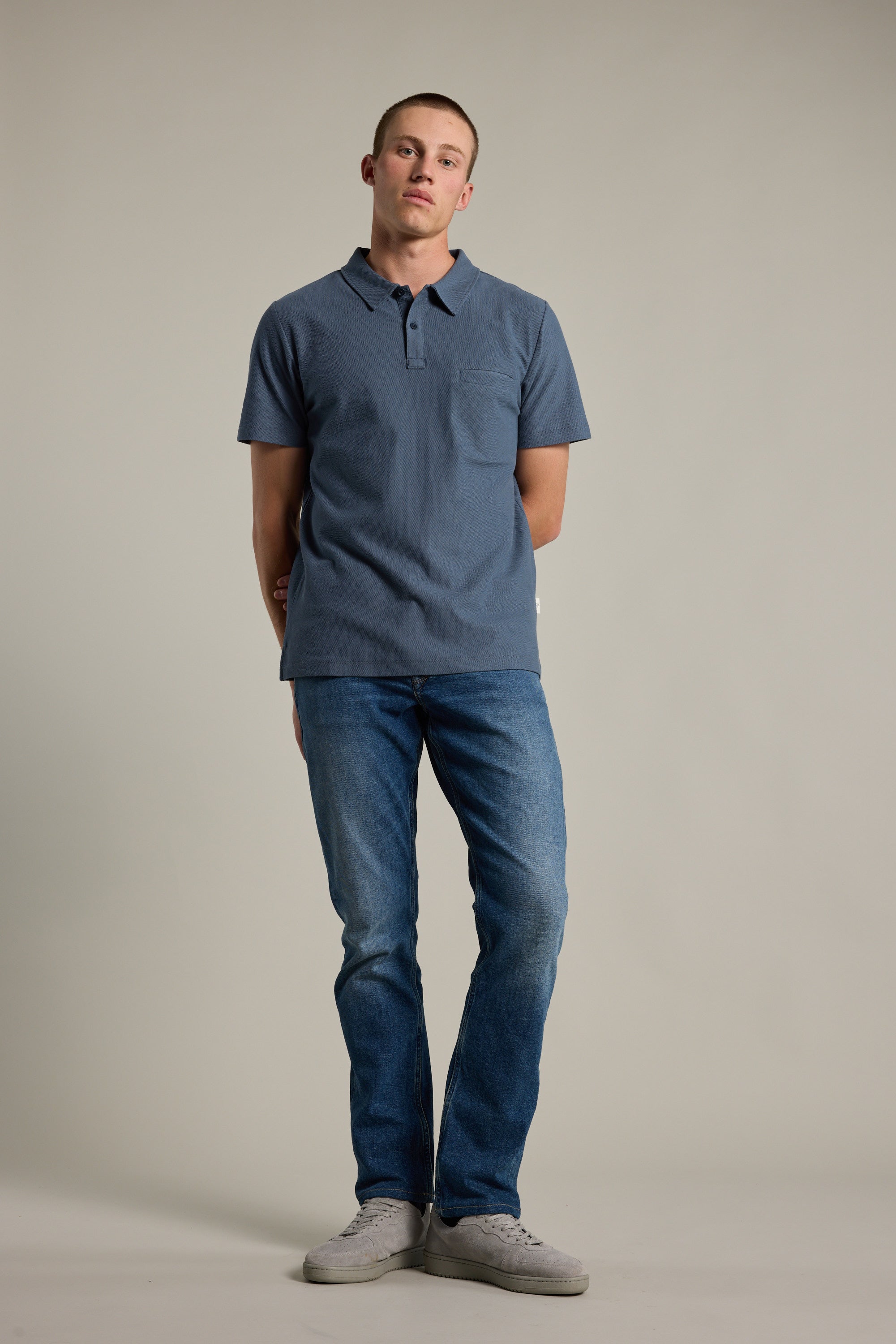 A person stands against a plain background, wearing a blue Barkers Canal Polo, blue jeans, and gray sneakers, with arms behind their back and a neutral expression.
