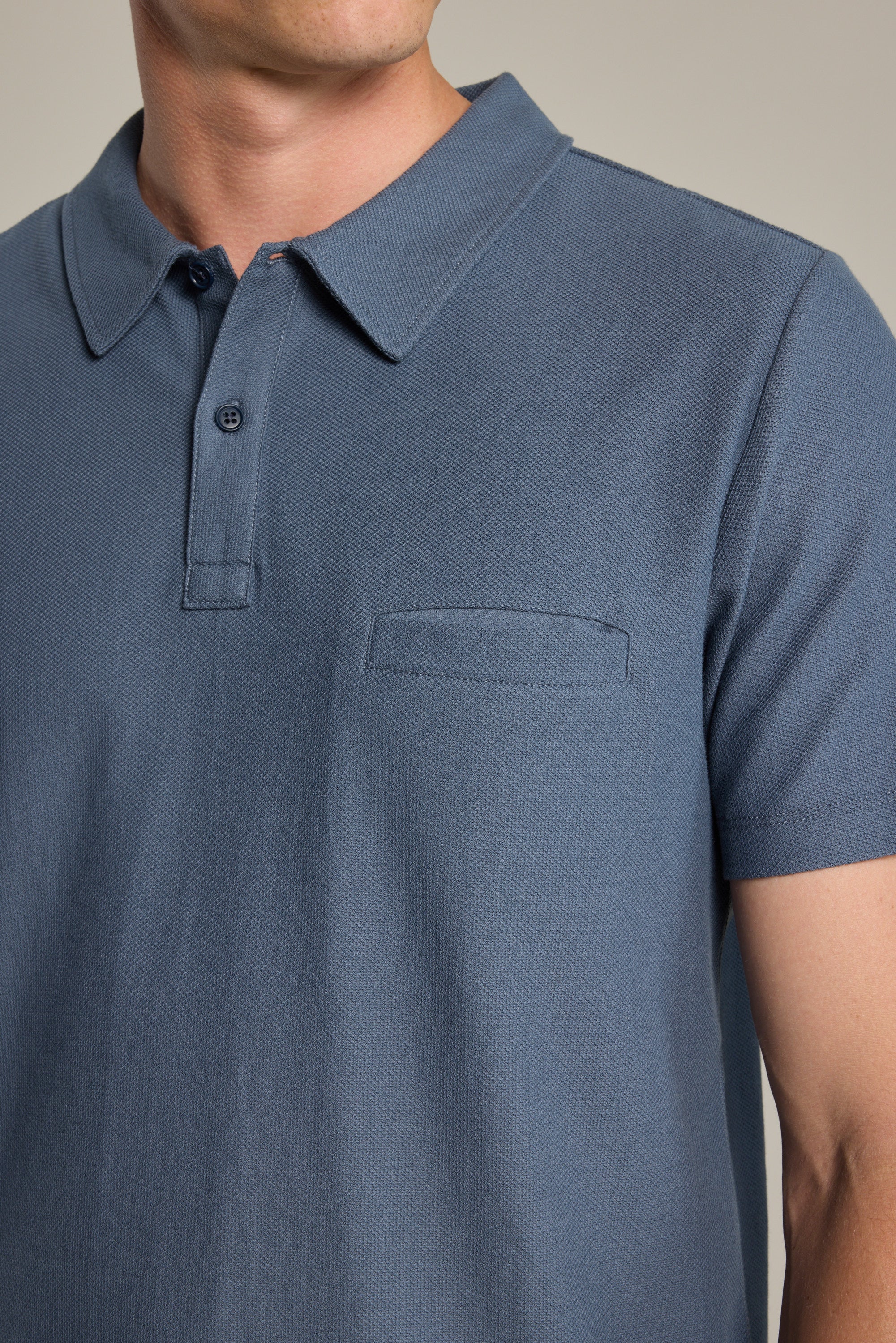 A person wearing the Barkers Canal Polo, a short-sleeved blue Supima cotton shirt with a button placket and small horizontal chest pocket, photographed from shoulders to waist.