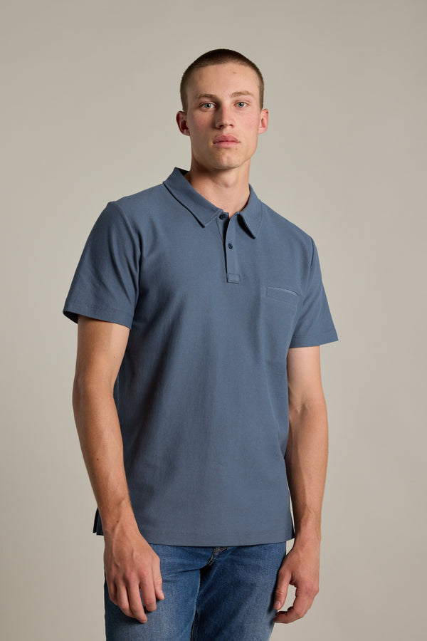 A young man with short hair stands against a plain background, wearing Barkers' Canal Polo and blue jeans, looking directly at the camera with a neutral expression.