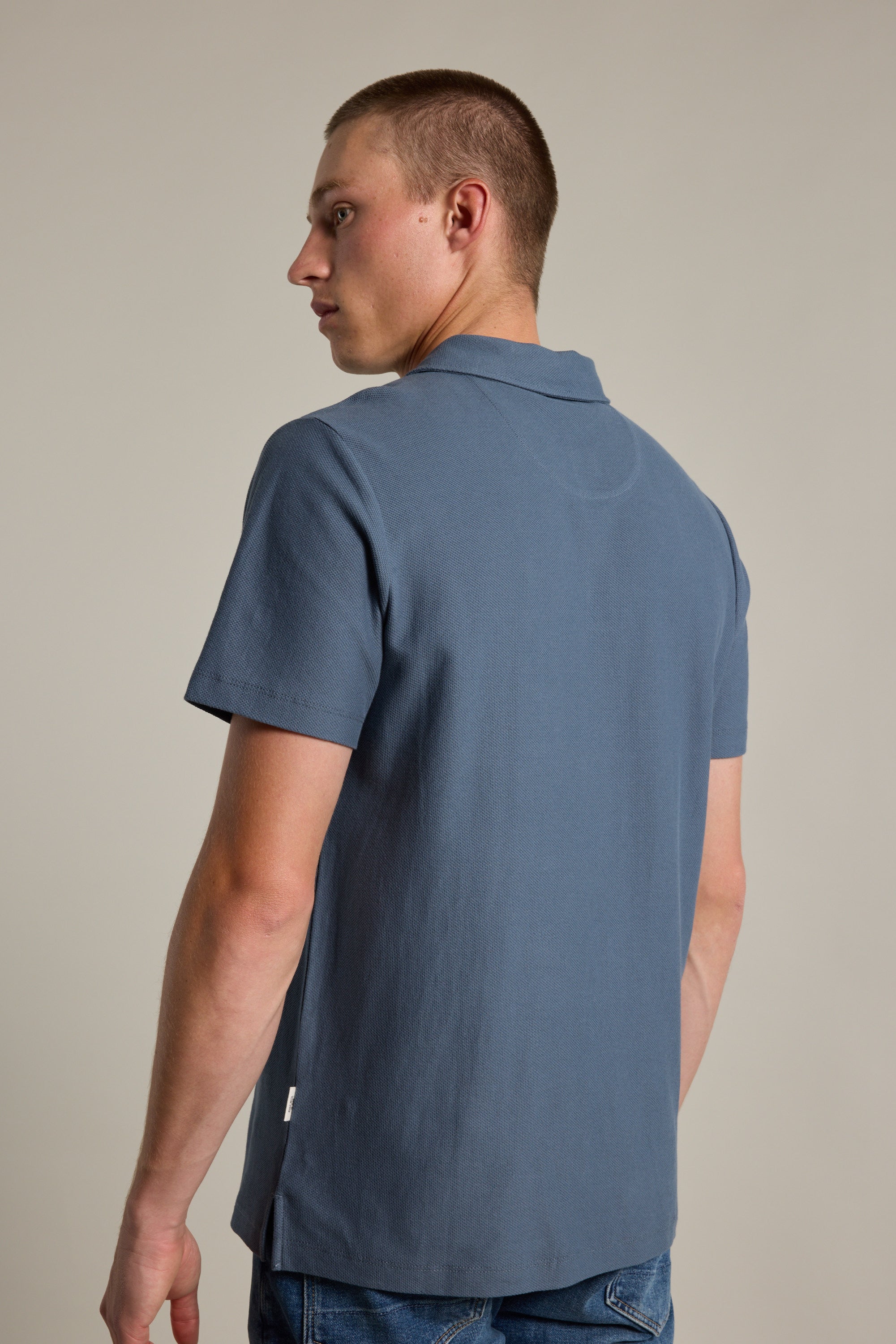 A person with short hair stands facing away, glancing over their left shoulder against a plain light background, modeling the Barkers Canal Polo in blue.