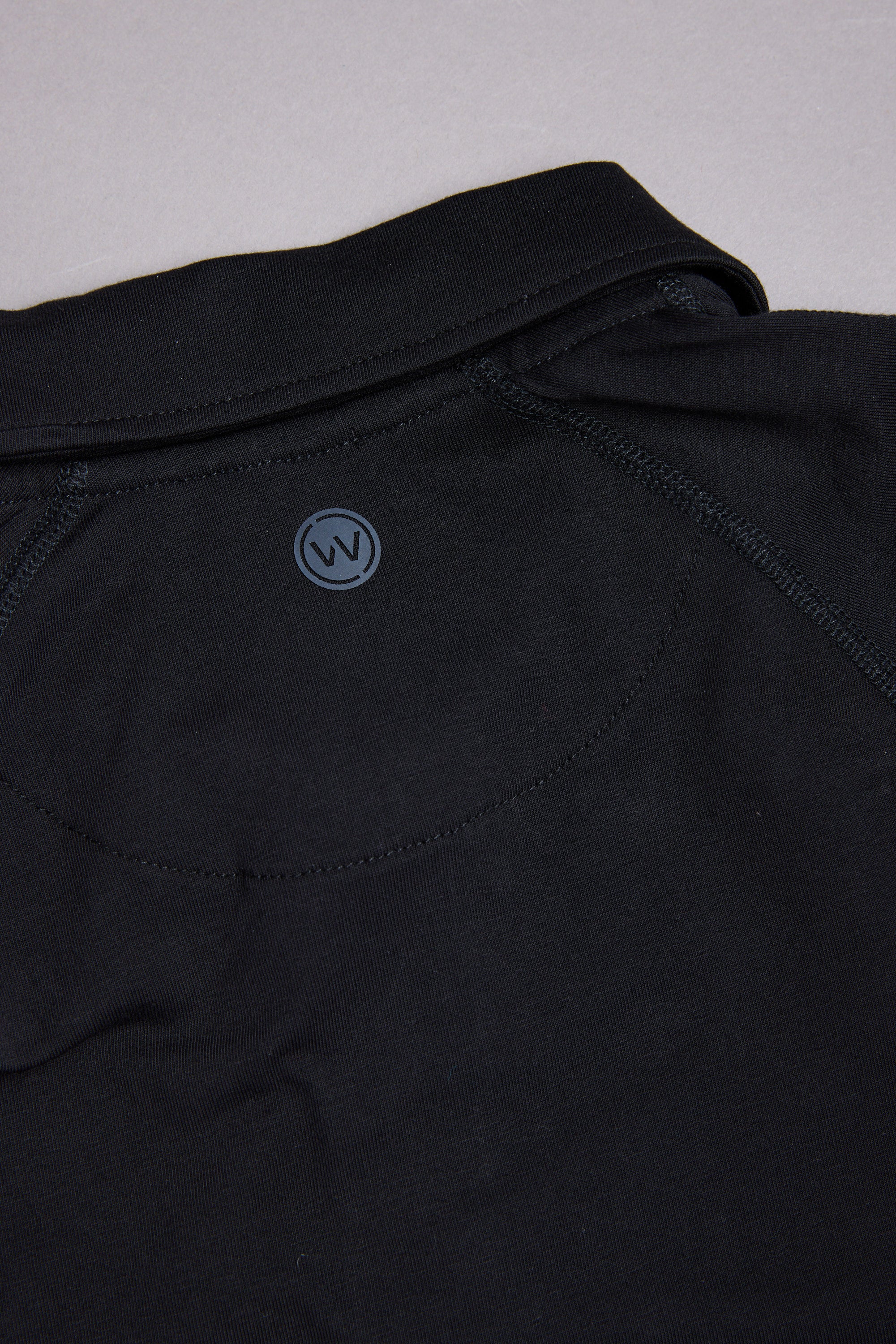 Close-up of the back of a black WearEver Outbound Polo, showcasing a small circular "W" logo near the neckline. Breathable cotton-blend fabric and stitching details highlight its lightweight stretch comfort.