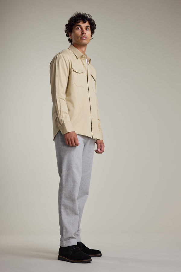 A man with curly dark hair stands in a neutral pose, wearing the Barkers Dunmore Pant in light gray—smart-casual pants made from responsibly sourced cotton—paired with a beige shirt and black shoes, against a plain light background.
