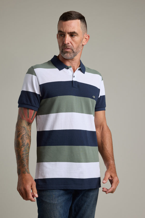 A man with short dark hair, a beard, and a colorful arm tattoo wears the Barkers Regent Pique Stripe Polo, featuring wide navy, green, and white stripes. He stands against a plain light background, looking to the side.