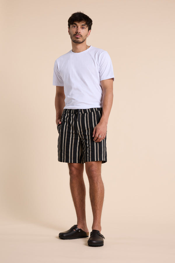 A man stands against a beige background wearing Barkers Lawton Stripe Shorts with an elastic waistband and a white T-shirt. He pairs the look with black slip-on shoes, his short dark hair neat, hands in his pockets.