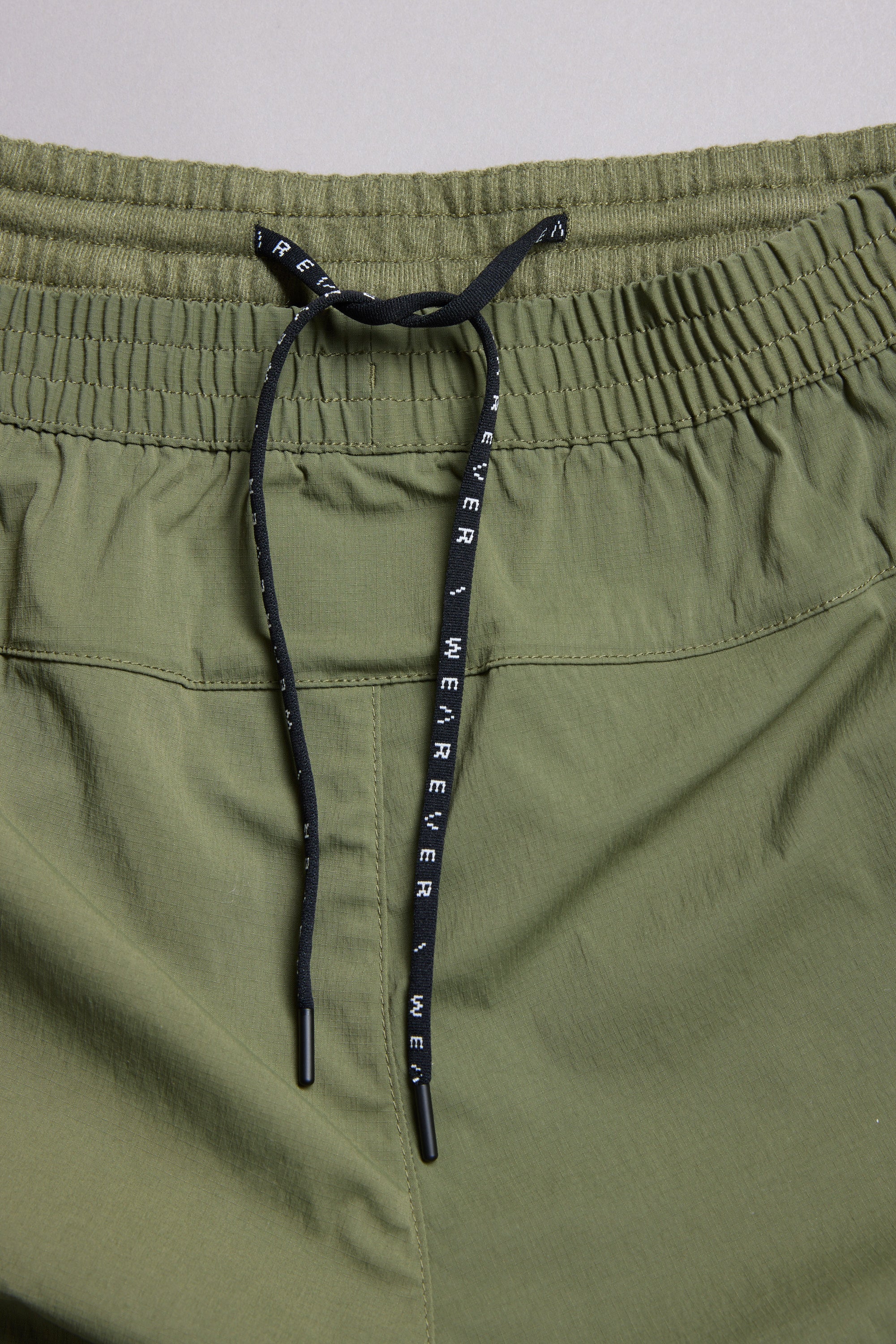 Close-up of WearEver Agile Short, olive green athletic shorts by WearEver with a 7-inch inseam, elastic waistband, and black drawstring bearing white WHEREVER text. The lightweight fabric has a slightly textured look.