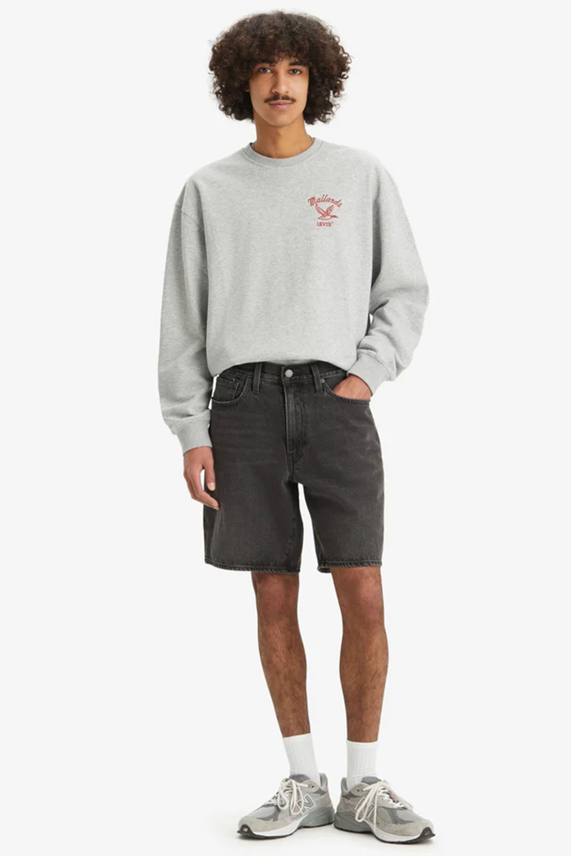 A person with curly hair wears a light gray sweatshirt, Levi's 468 Loose Short by Levis in black non-stretch denim, white socks, and gray sneakers, standing with one hand in their pocket against a plain white background.
