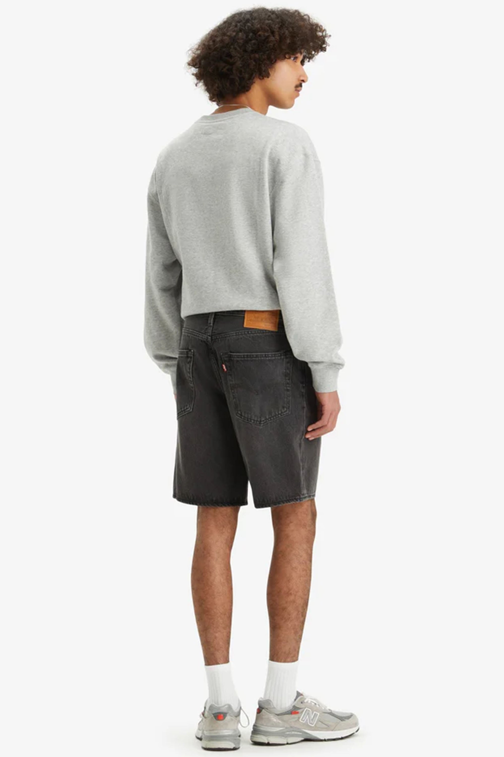 A person stands facing away, wearing a light gray sweatshirt, Levi’s 468 Loose Shorts in black non-stretch denim, white socks, and gray sneakers. The background is plain white.