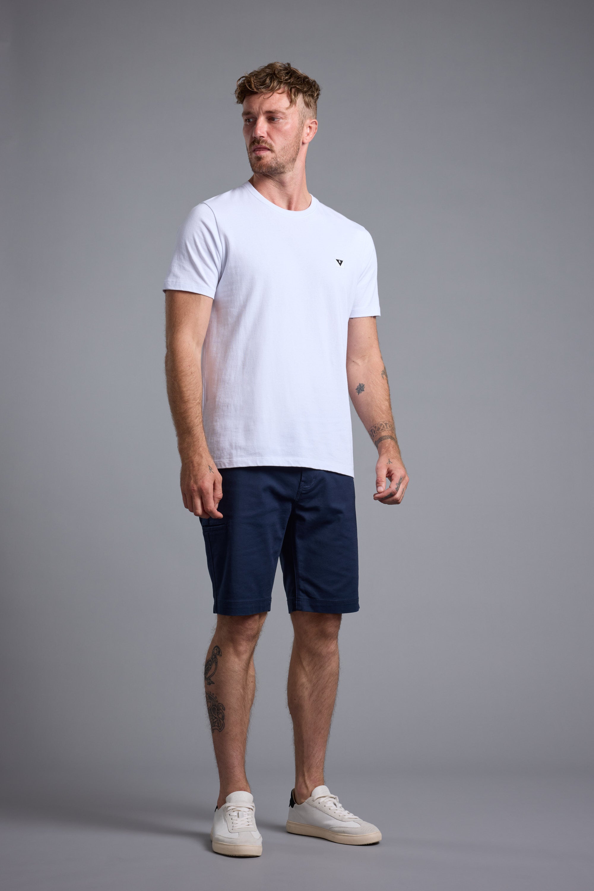 A man with tattoos on his left arm and leg stands against a gray background, wearing a white t-shirt, Barkers Anvil 5Pkt Short in navy blue with four-way stretch, and white sneakers. He looks slightly to his left.