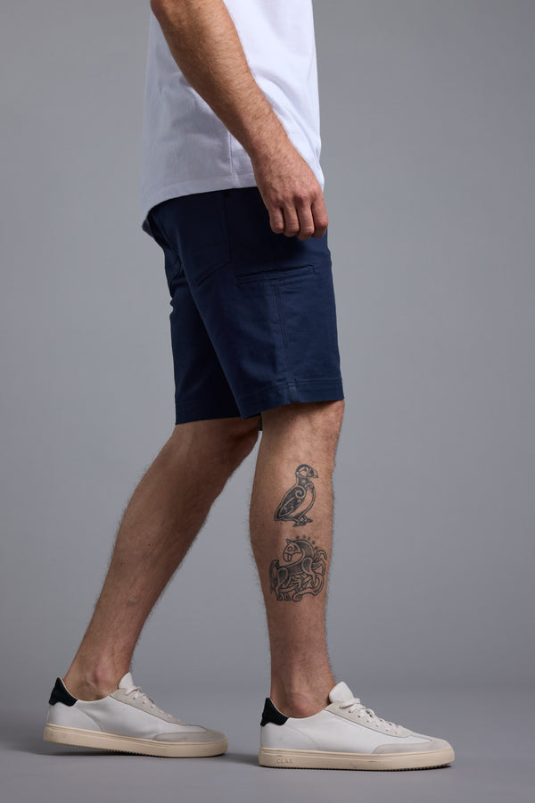 A person stands sideways in a white t-shirt, Barkers Anvil 5Pkt Shorts in navy, and white sneakers. Tattoos are visible on the left calf against a plain gray background.
