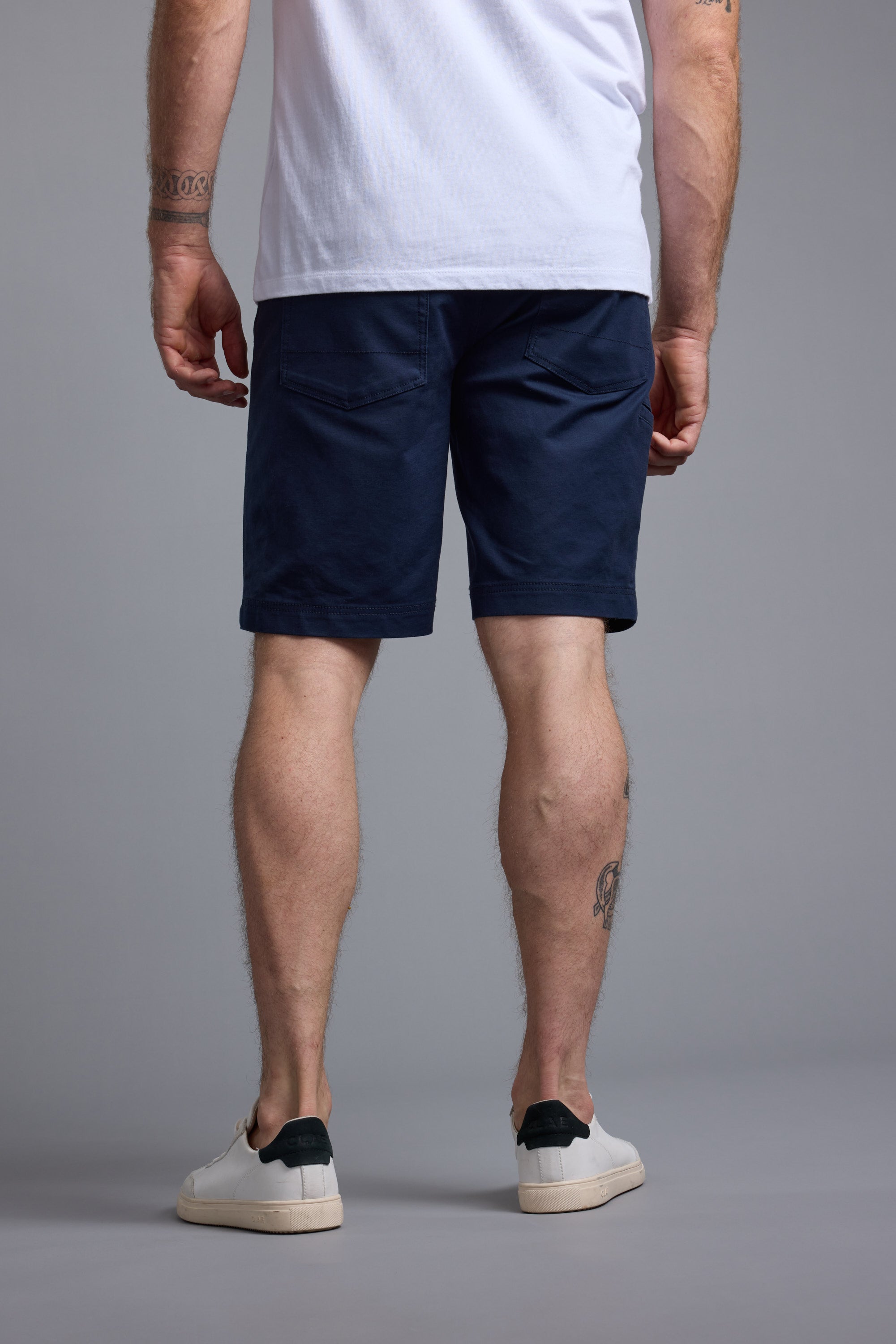 A man faces away from the camera, wearing a white T-shirt, white sneakers, and navy Barkers Anvil 5Pkt Shorts made from a durable cotton blend. Tattoos are visible on his arms and left calf. The backdrop is plain gray.