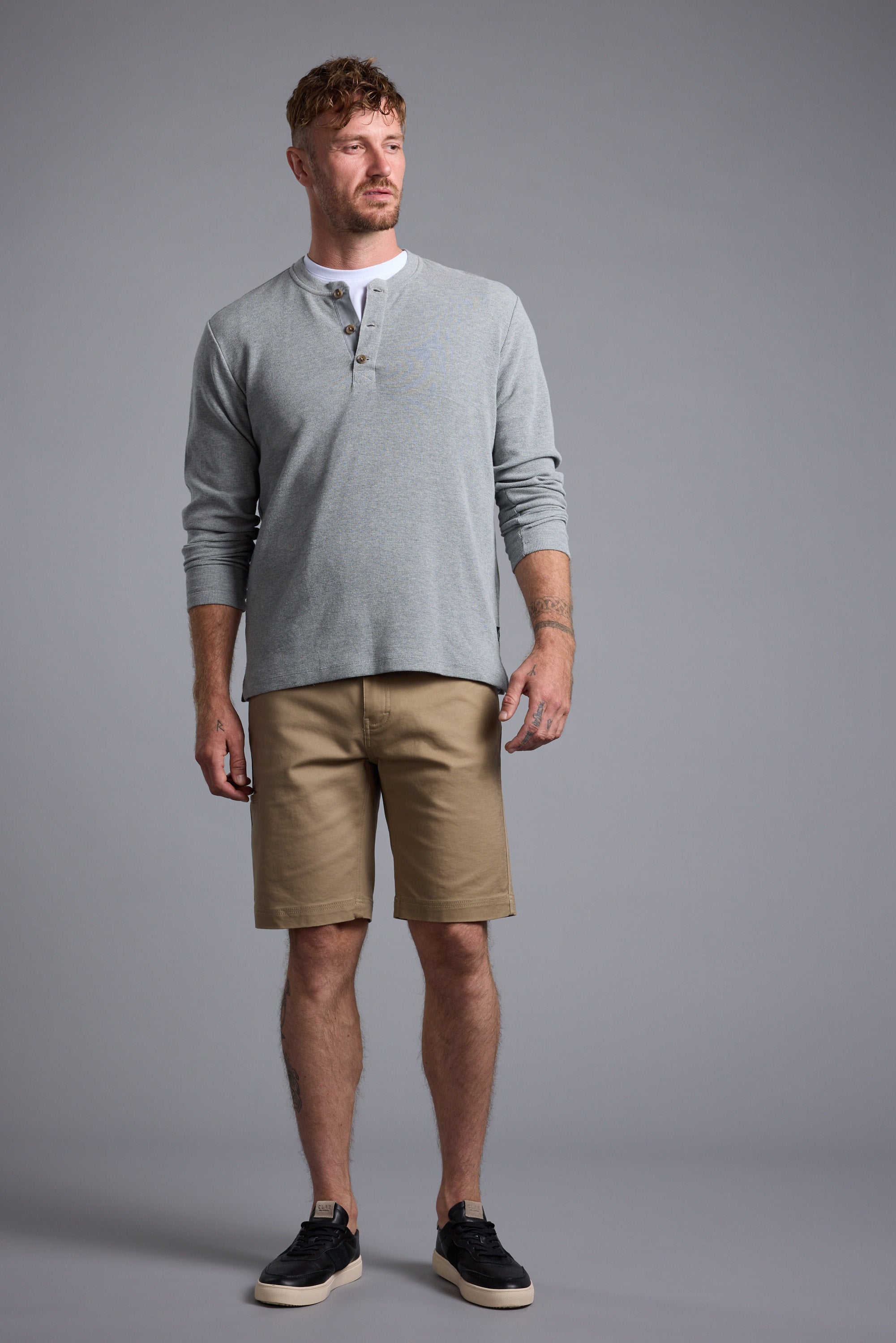 A man with short hair and a beard stands against a gray background, wearing a light gray long-sleeve henley shirt, Barkers Anvil 5Pkt Short in tan with an elasticated waistband, and black sneakers with white soles. He looks slightly left, expression neutral.