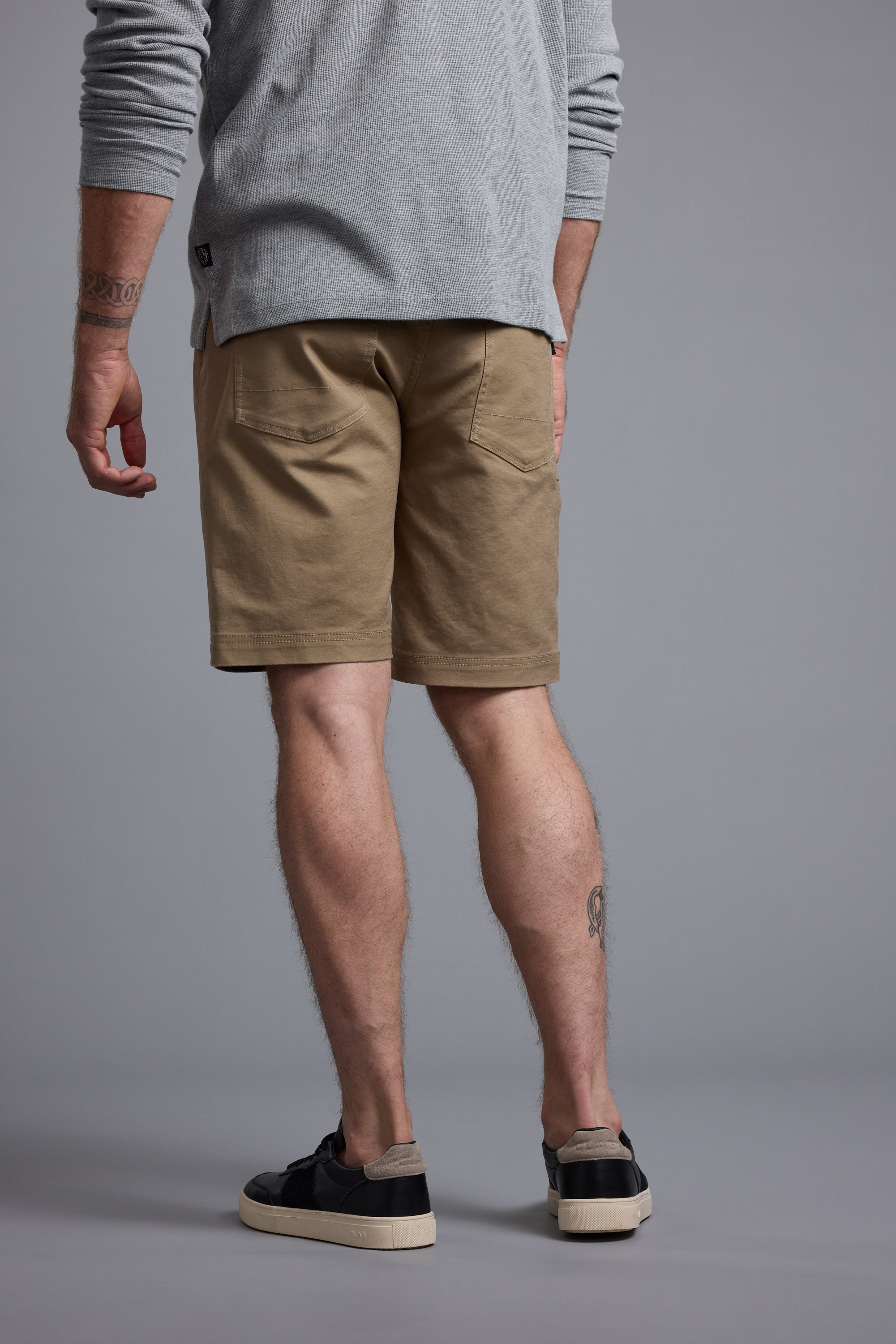A person faces away, wearing a light gray long-sleeve shirt and the Barkers Anvil 5Pkt Short with an elasticated waistband. Black sneakers complete the look, revealing tattooed lower legs against a plain gray background.