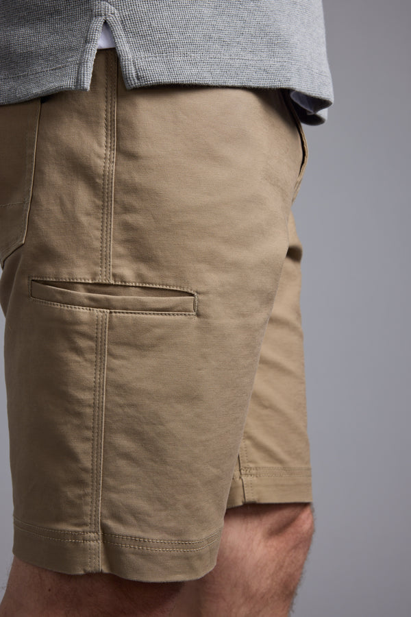 A close-up shows a person wearing Barkers Anvil 5Pkt Shorts in tan with an elastic waistband, side pocket, stitched details, and four-way stretch fabric. Only the lower torso and upper legs are visible.