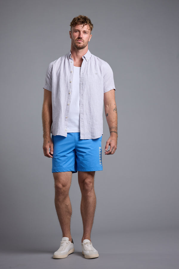 A man with short hair and a neutral expression stands against a plain gray backdrop, wearing a light short-sleeve shirt over a white tank, bright blue Barkers Weekend Shorts by Barkers made from recycled polyester, and white sneakers.