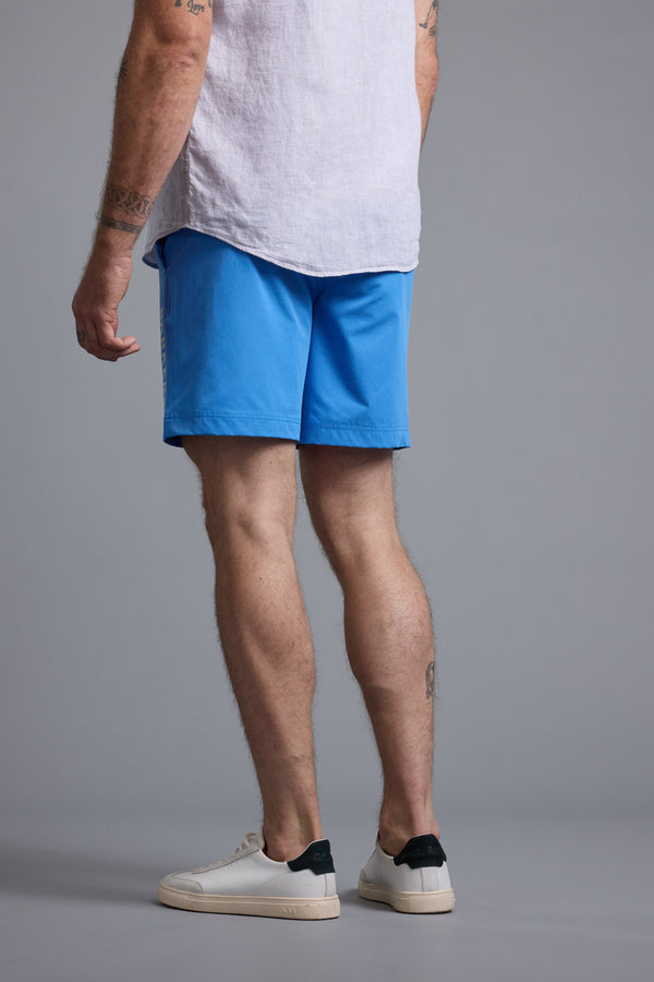 A man stands with his back to the camera, wearing a light shirt and bright blue Barkers Weekend Shorts by Barkers, made from recycled polyester, paired with white sneakers featuring dark heel accents against a plain gray background.