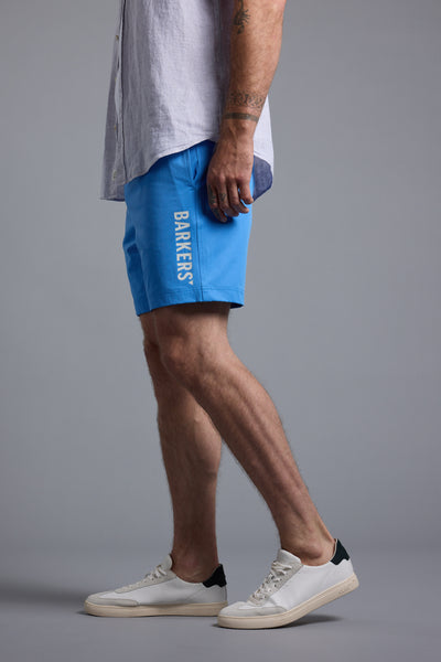 A person stands sideways against a plain grey background wearing a light grey short-sleeve shirt, Barkers Weekend Short in bright blue with BARKERS branding on the side, and white sneakers with black heel accents.
