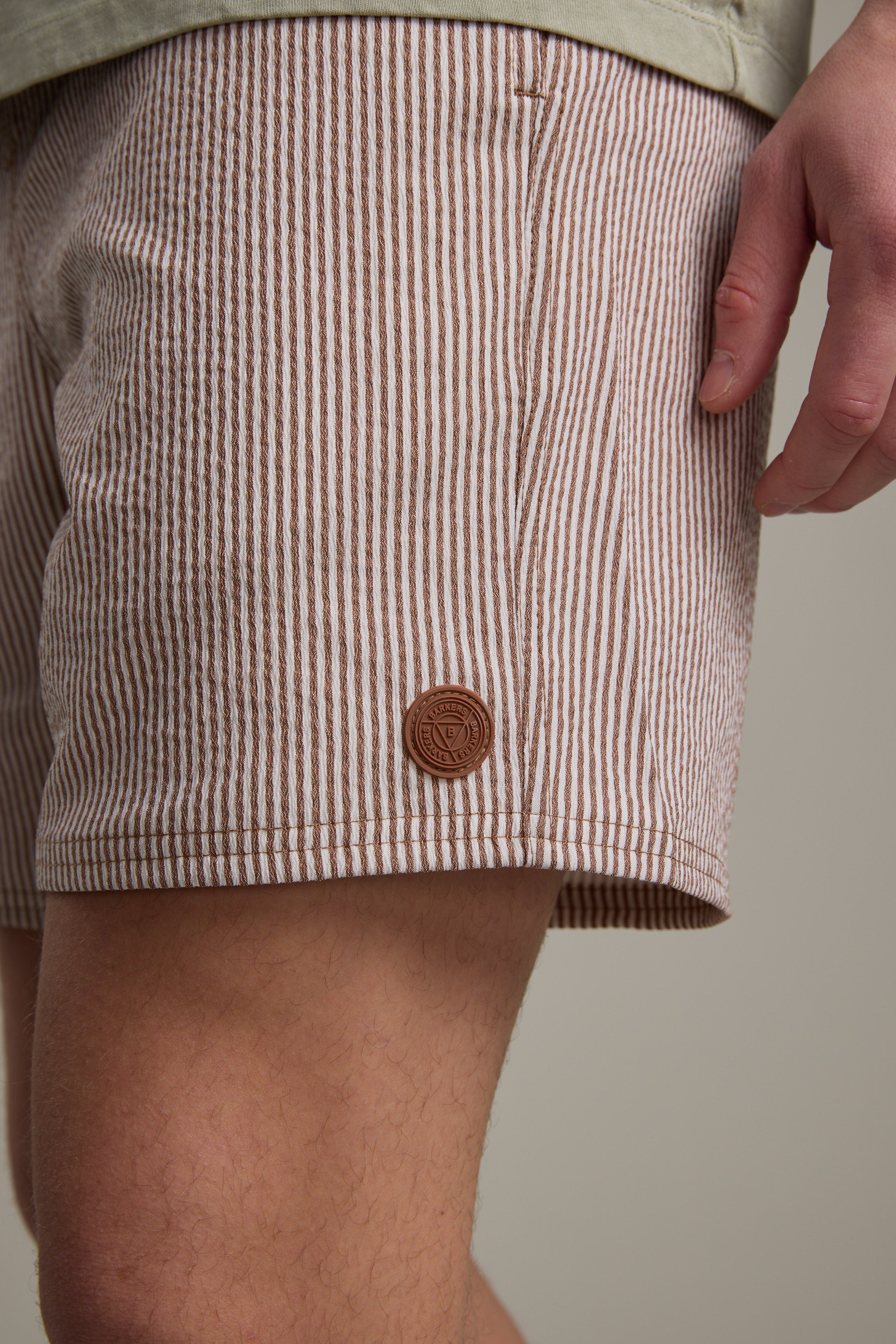 A close-up of a person wearing Barkers Dehaven Stripe Volley shorts with beige and white seersucker stripes and a small brown patch near the hem, their right hand resting at their side.
