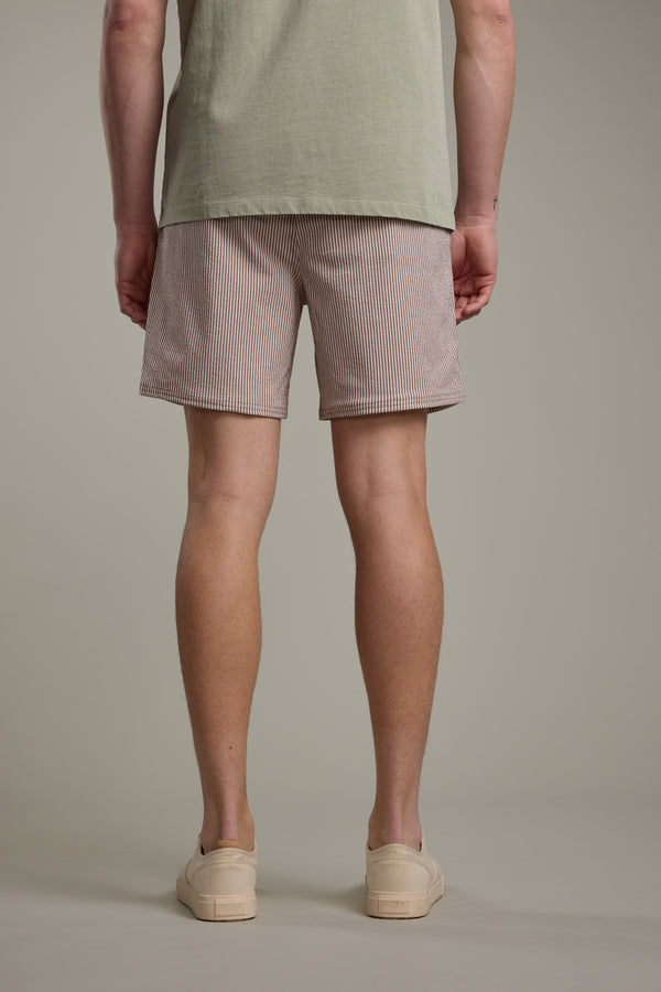 A person stands with their back to the camera, wearing a light green t-shirt, Barkers Dehaven Stripe Volley red and white seersucker shorts, and beige slip-on shoes against a plain background.