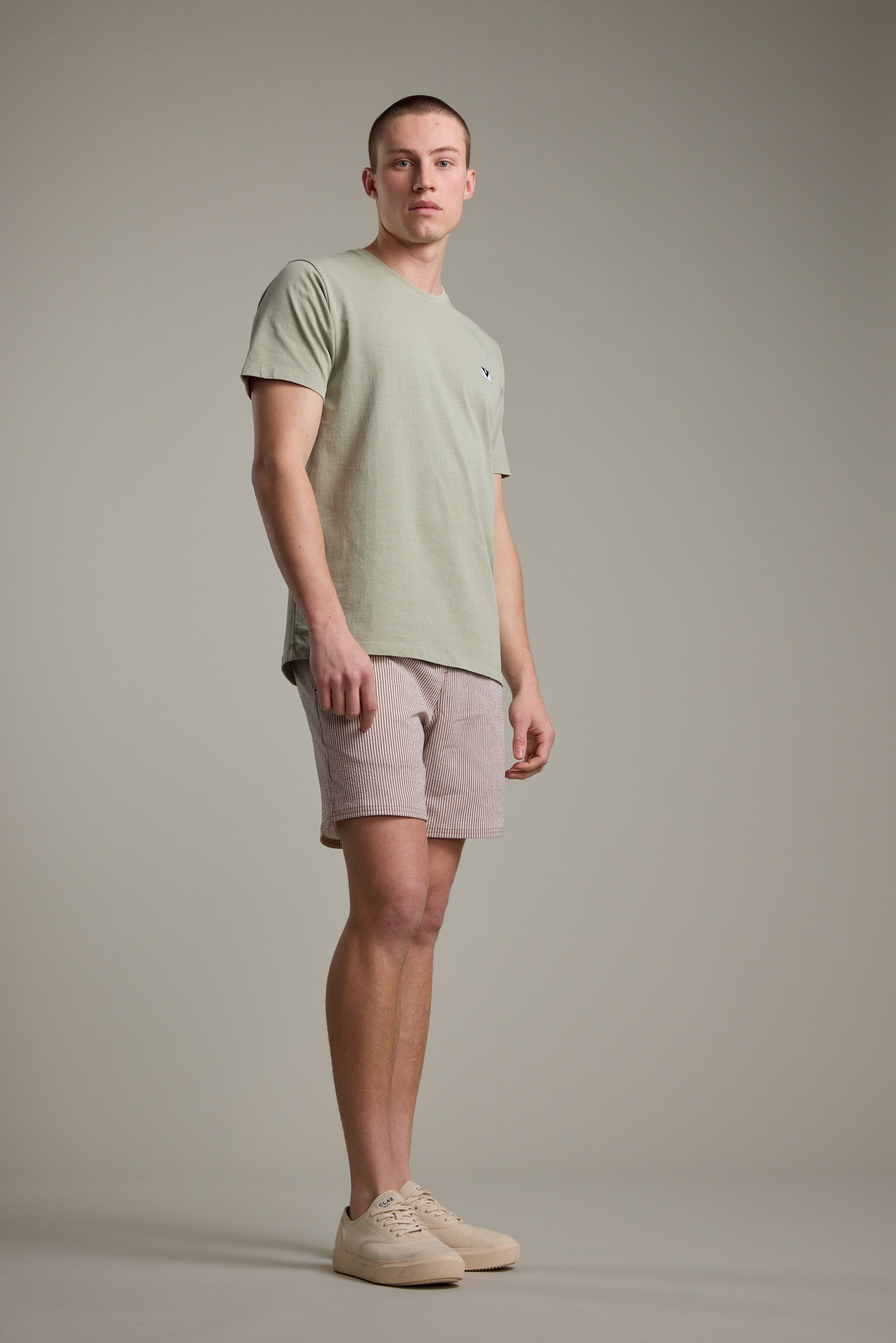 A man stands against a plain, light gray background wearing Barkers’ Dehaven Stripe Volley seersucker shorts in recycled polyester, a light green T-shirt, and beige sneakers. He faces slightly sideways with a neutral expression.