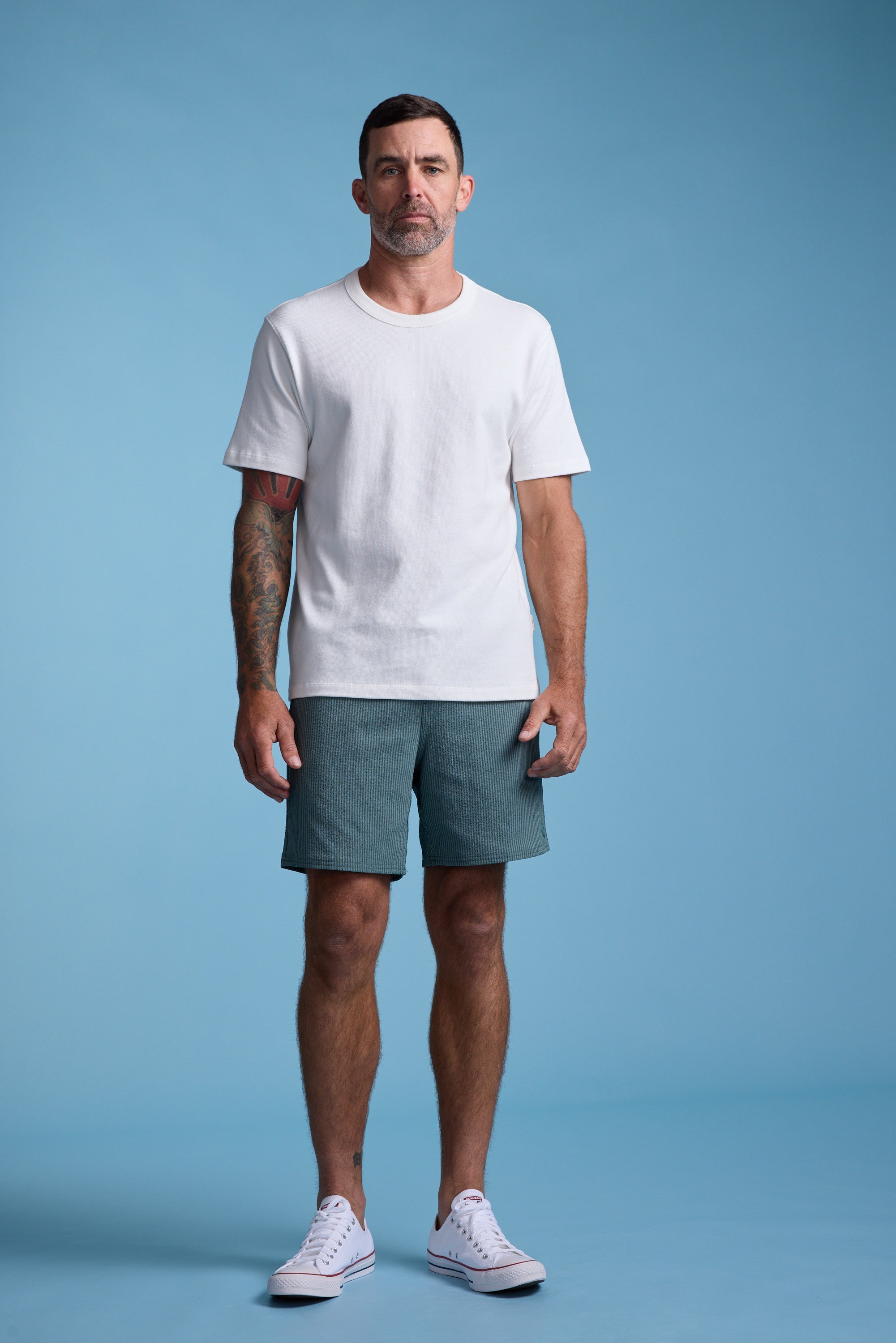A man with short dark hair and tattoos on his left arm stands against a blue background, wearing a white T-shirt, Barkers Dehaven Stripe Volley quick-drying swim trunks in green, and white sneakers.