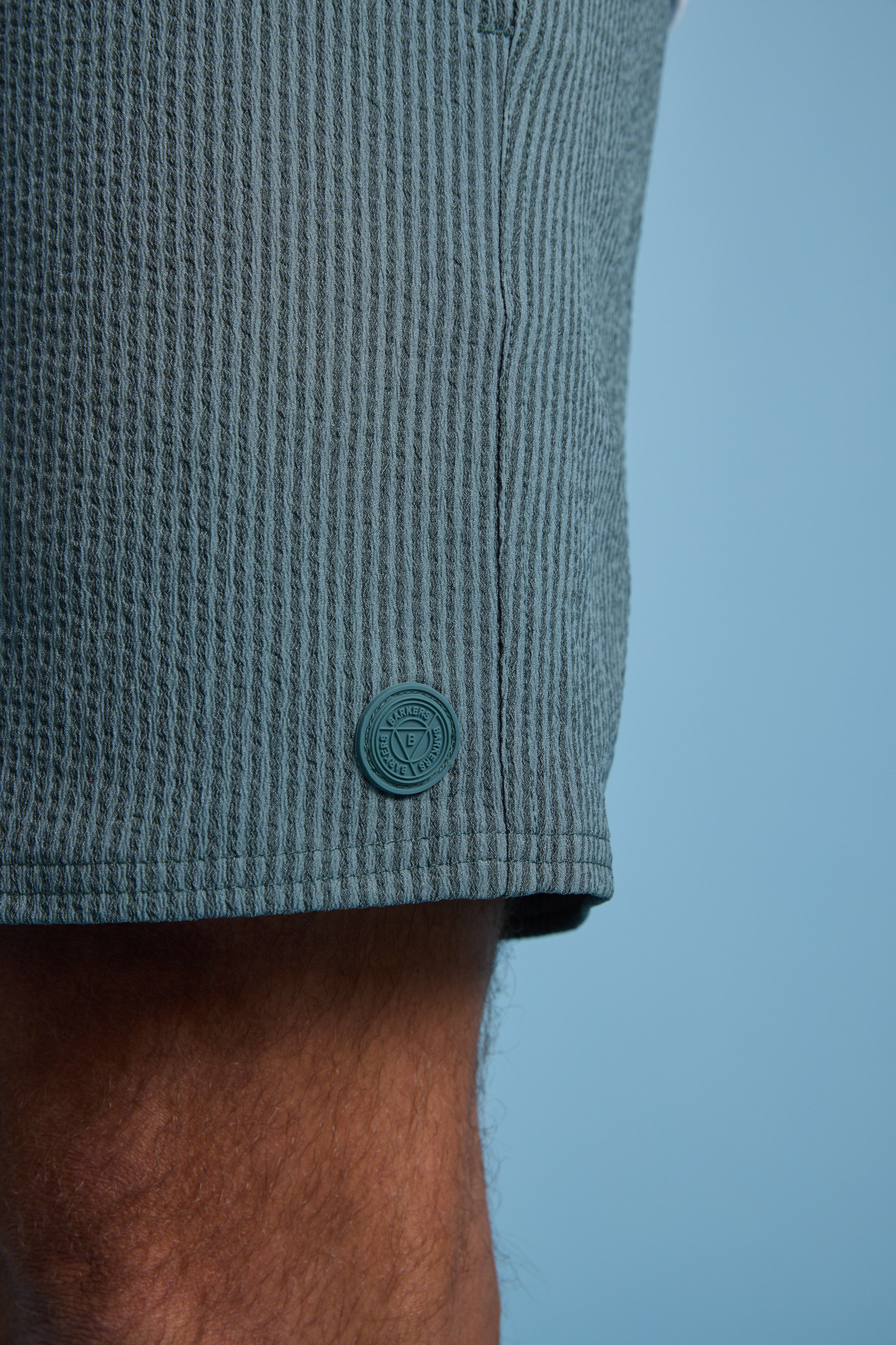 Close-up of a person's upper leg wearing Barkers Dehaven Stripe Volley shorts in textured, light greenish-blue seersucker with a matching round button and small emblem by the hem, set against a plain light blue background.