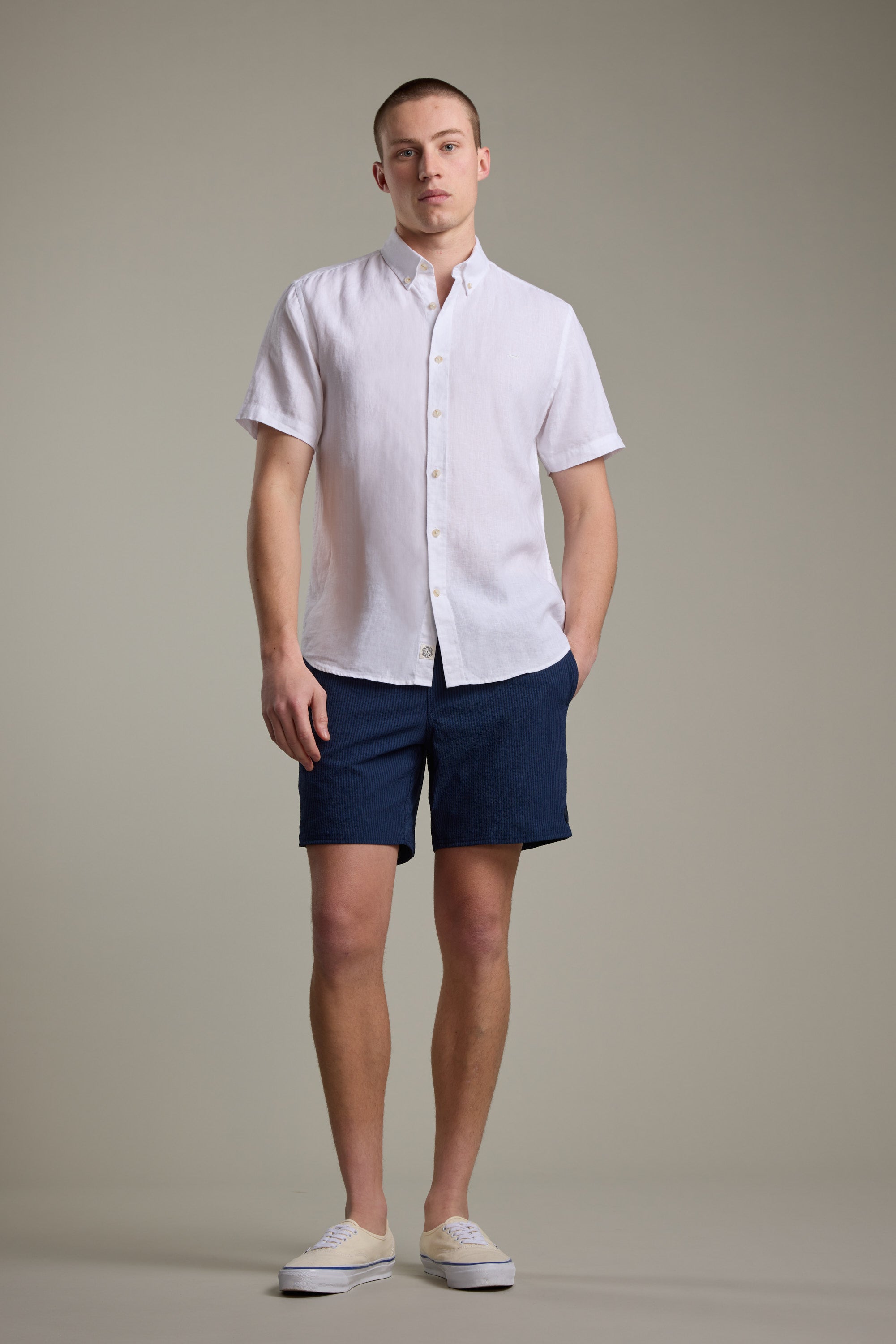 A man stands facing forward with one hand in his pocket, wearing Barkers Dehaven Stripe Volley navy seersucker shorts, a white short-sleeve button-up shirt, and beige sneakers. He has a neutral expression against a plain, light background.