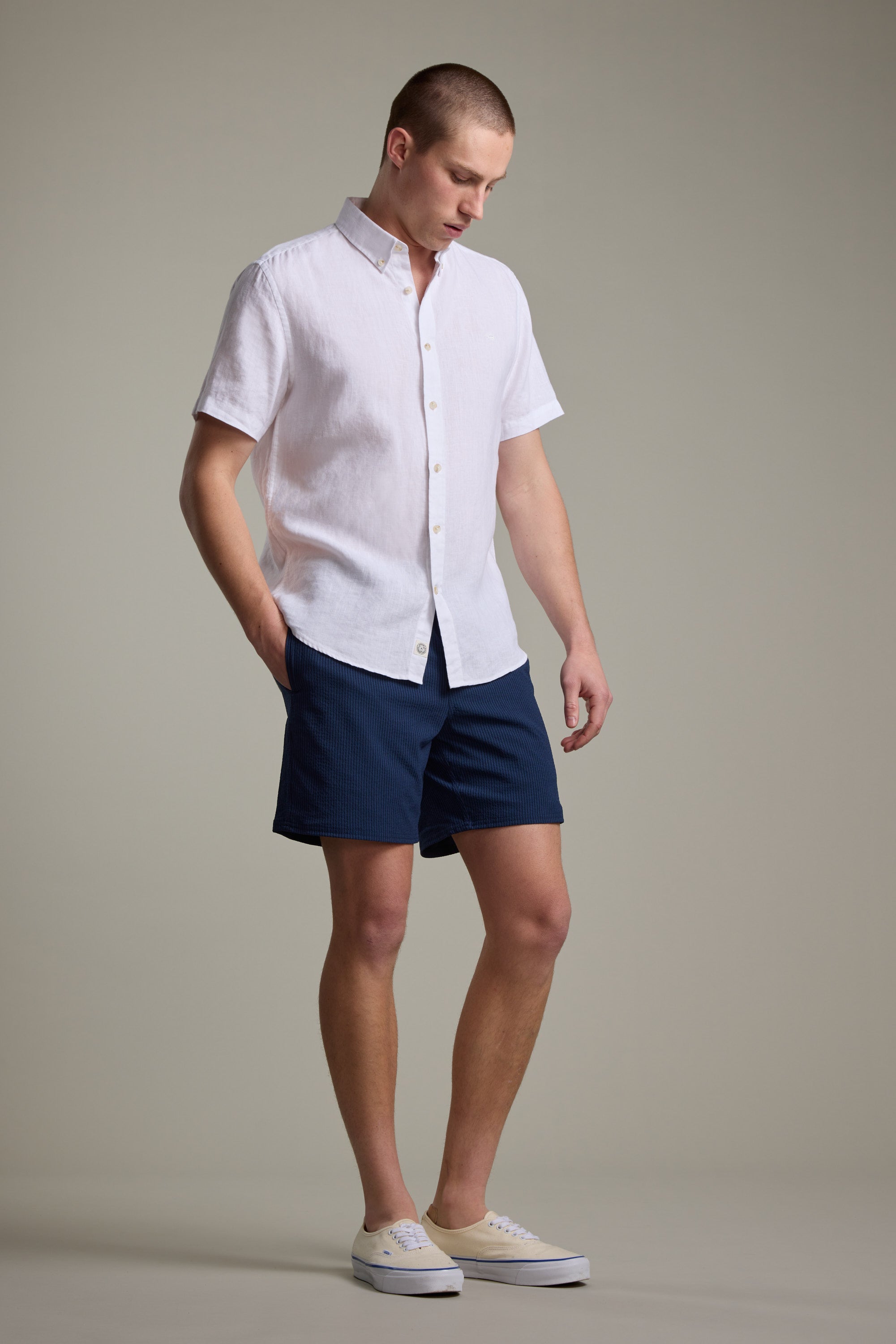A person stands against a plain background wearing Barkers' Dehaven Stripe Volley shorts, a short-sleeve white button-up shirt, and beige slip-on shoes, looking down with hands relaxed at their sides.