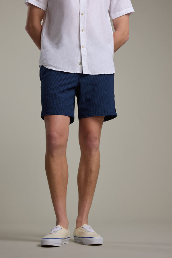A person stands with hands behind their back, wearing Barkers’ Dehaven Stripe Volley navy blue seersucker shorts, a white short-sleeve button-up shirt, and white sneakers. The background is plain and the person's face is not visible.
