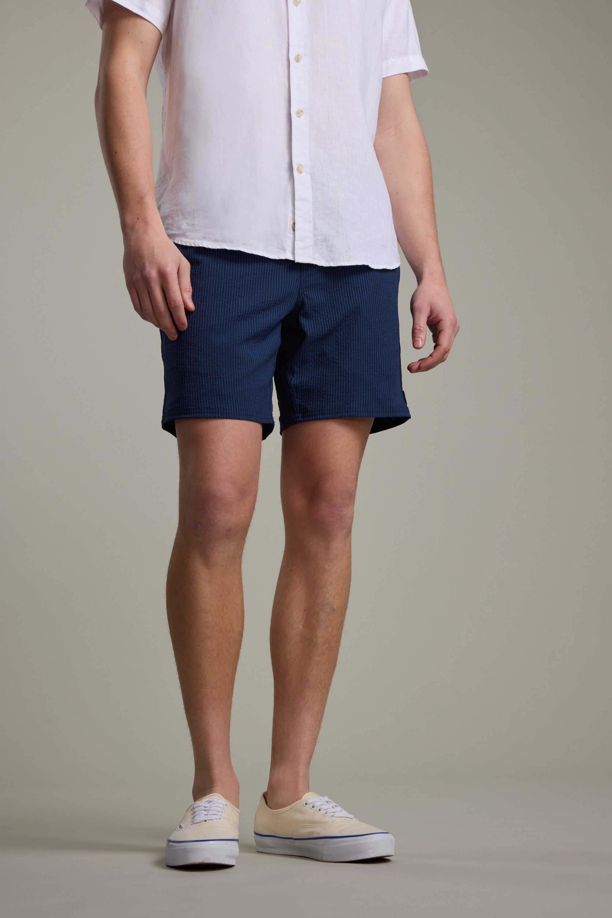 A person is standing against a plain background, wearing Barkers’ Dehaven Stripe Volley shorts in dark blue, a white short-sleeve button-up shirt, and white sneakers. The head and upper chest are not visible.