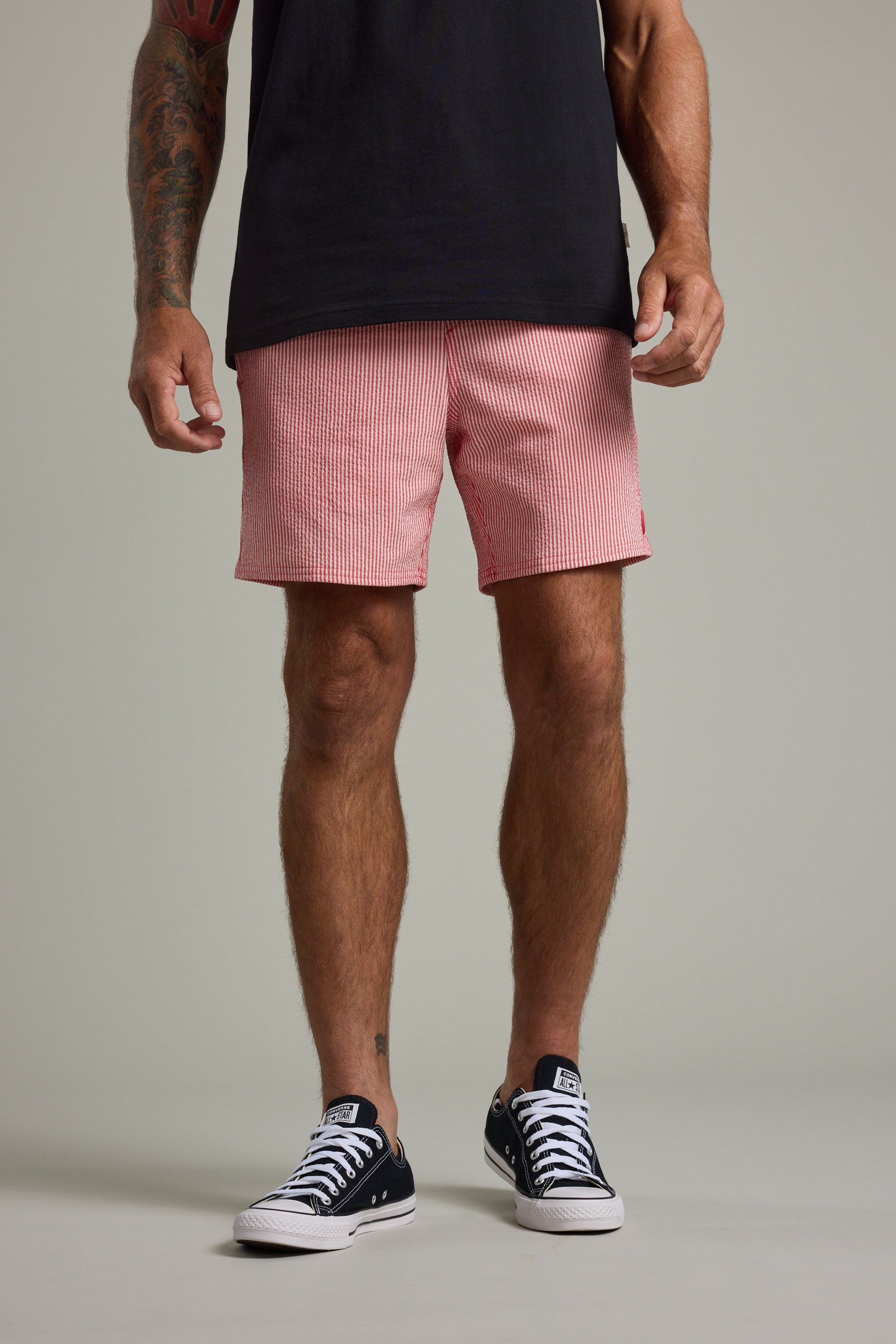 A person stands against a plain background, wearing black low-top sneakers, a black t-shirt, and Barkers Dehaven Stripe Volley quick-drying seersucker shorts made from recycled polyester. Only their tattooed shoulders-down body is visible.