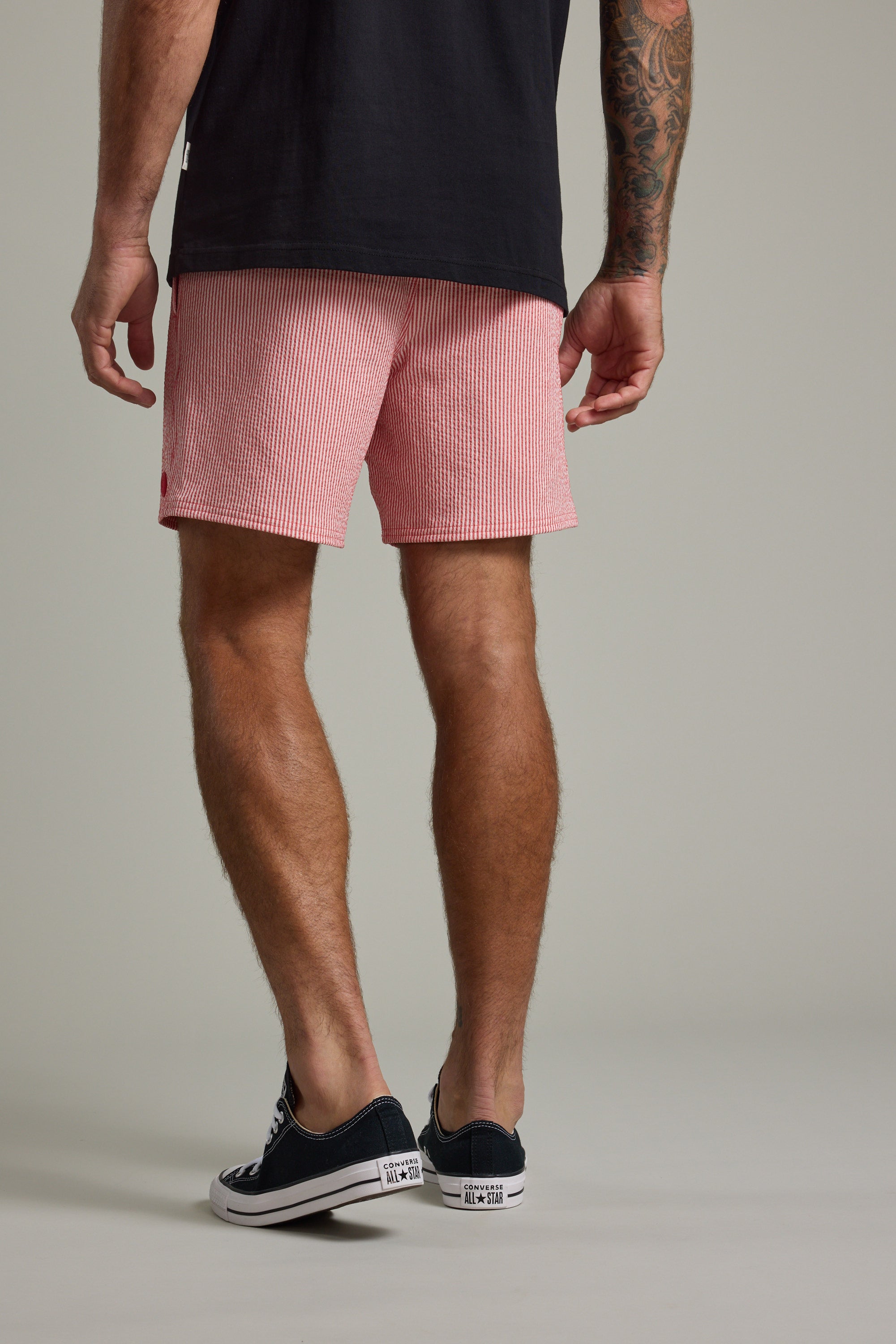 A man faces away in a black T-shirt, Barkers Dehaven Stripe Volley seersucker shorts, and black low-top sneakers, revealing a tattoo on his right forearm. The backdrop is plain and neutral.